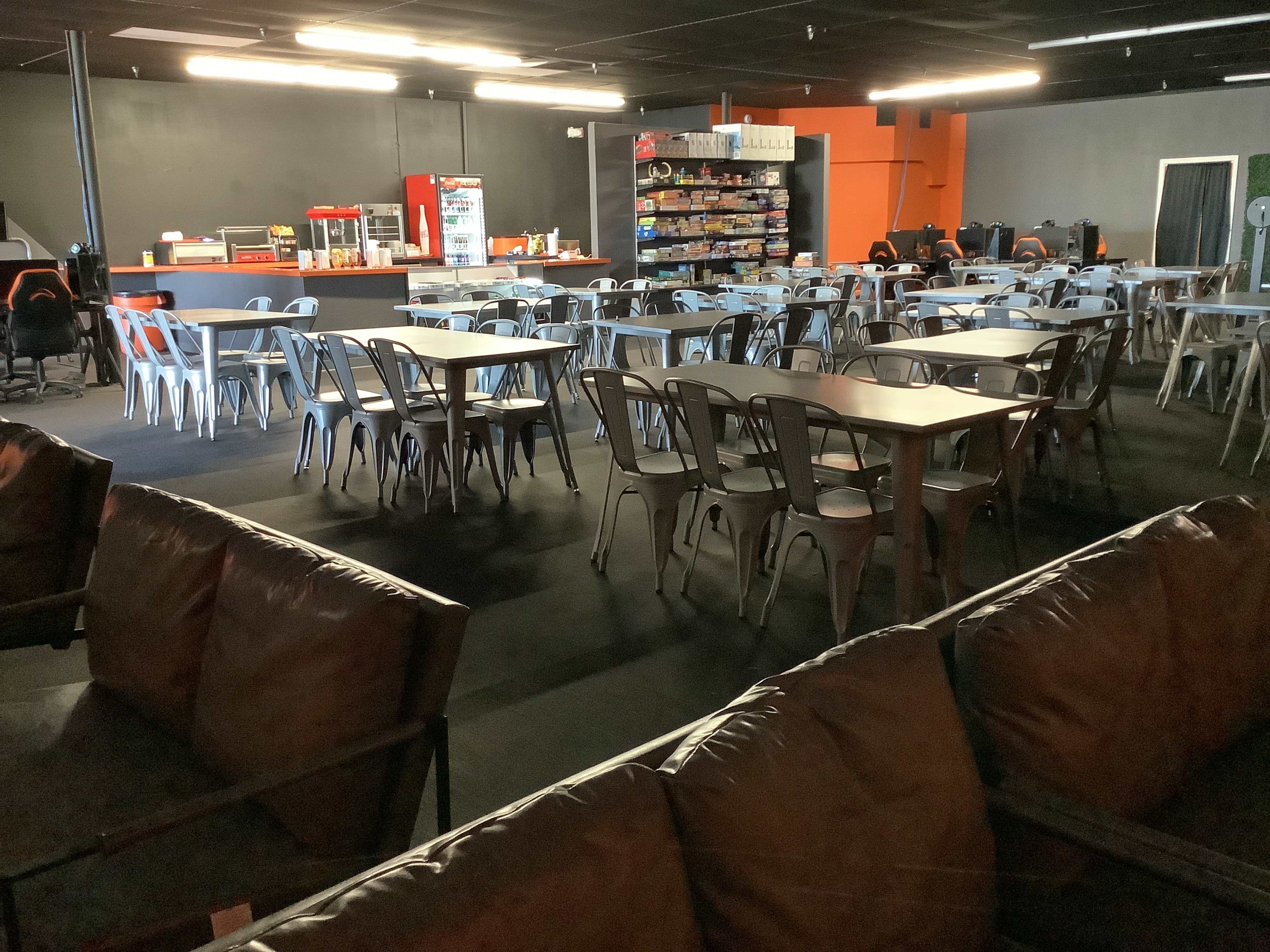 The image shows a spacious indoor area with rows of tables and chairs, a snack counter, and a lounge section with brown couches.