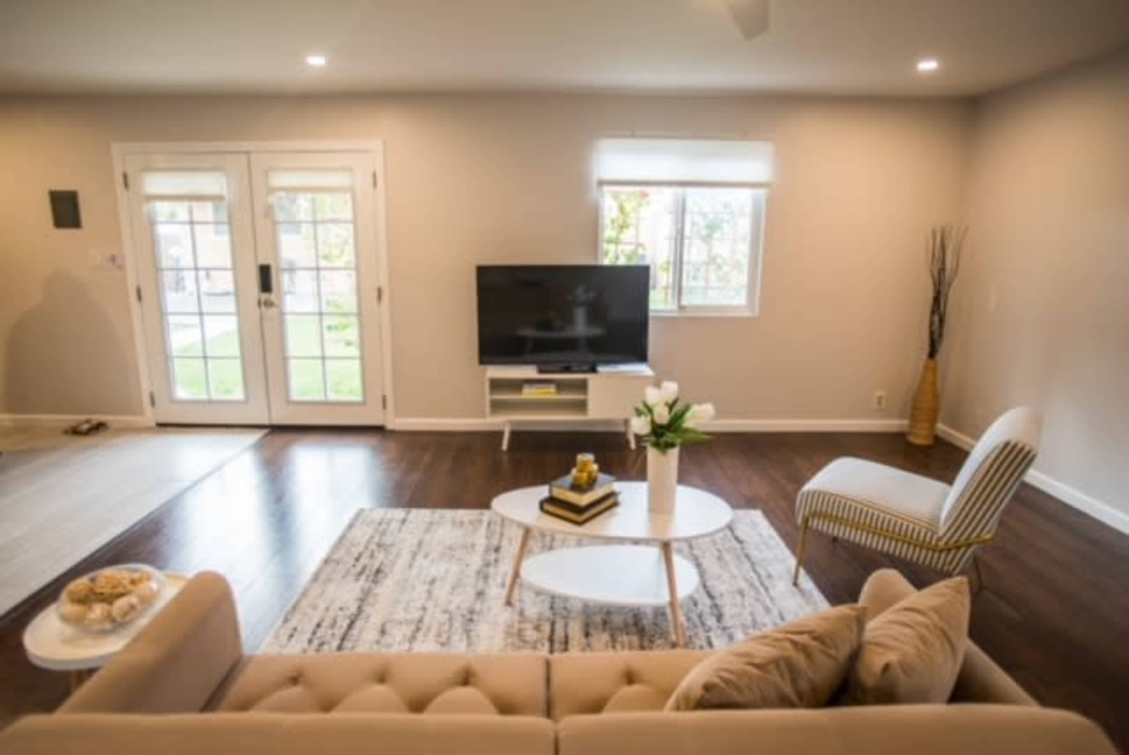 The living room features a beige sofa, a coffee table with flowers, a television on a stand, and a striped chair beside large windows and a set of double doors.