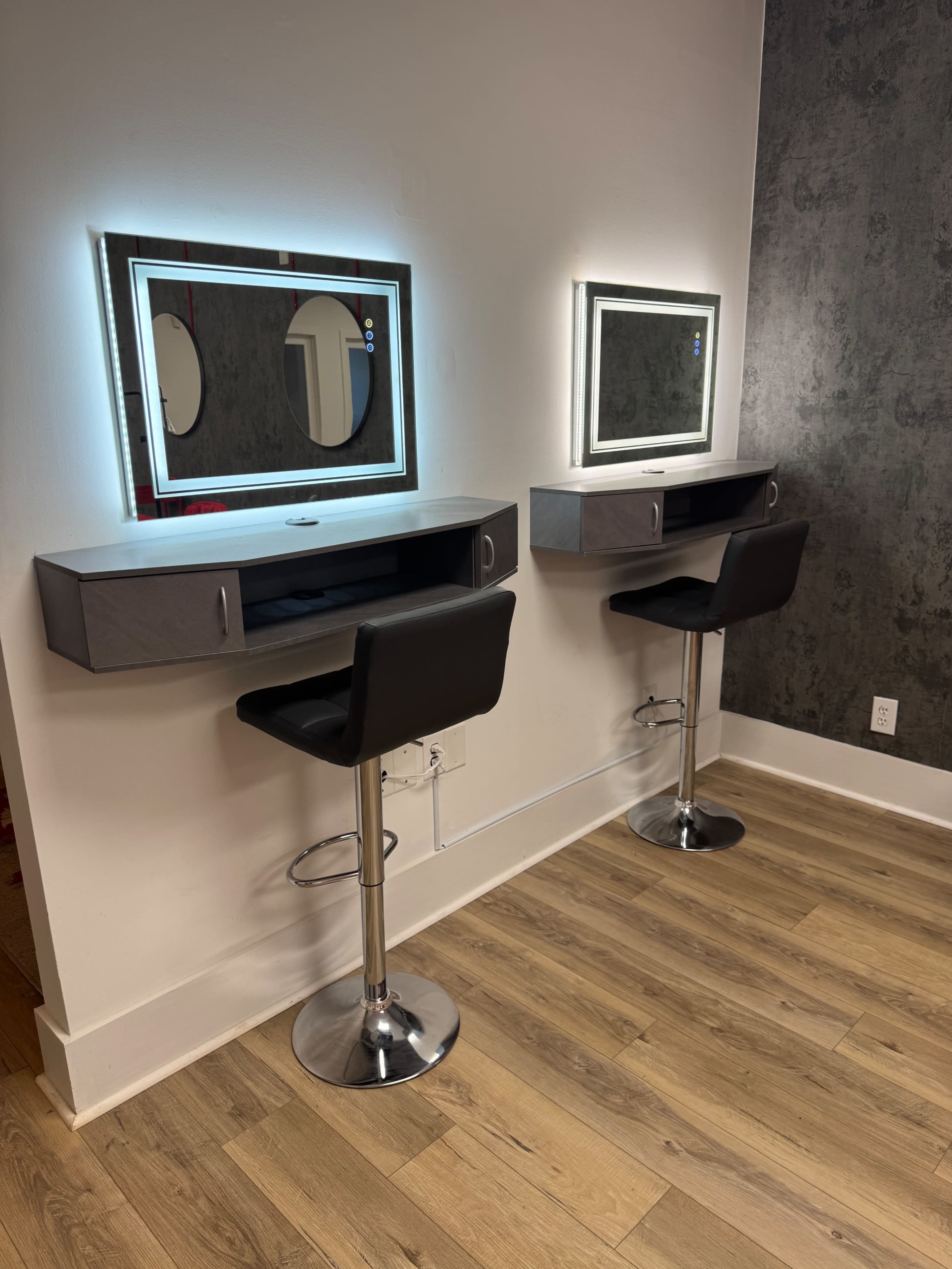 The image shows two modern vanity stations with illuminated mirrors and black stools against a textured wall.