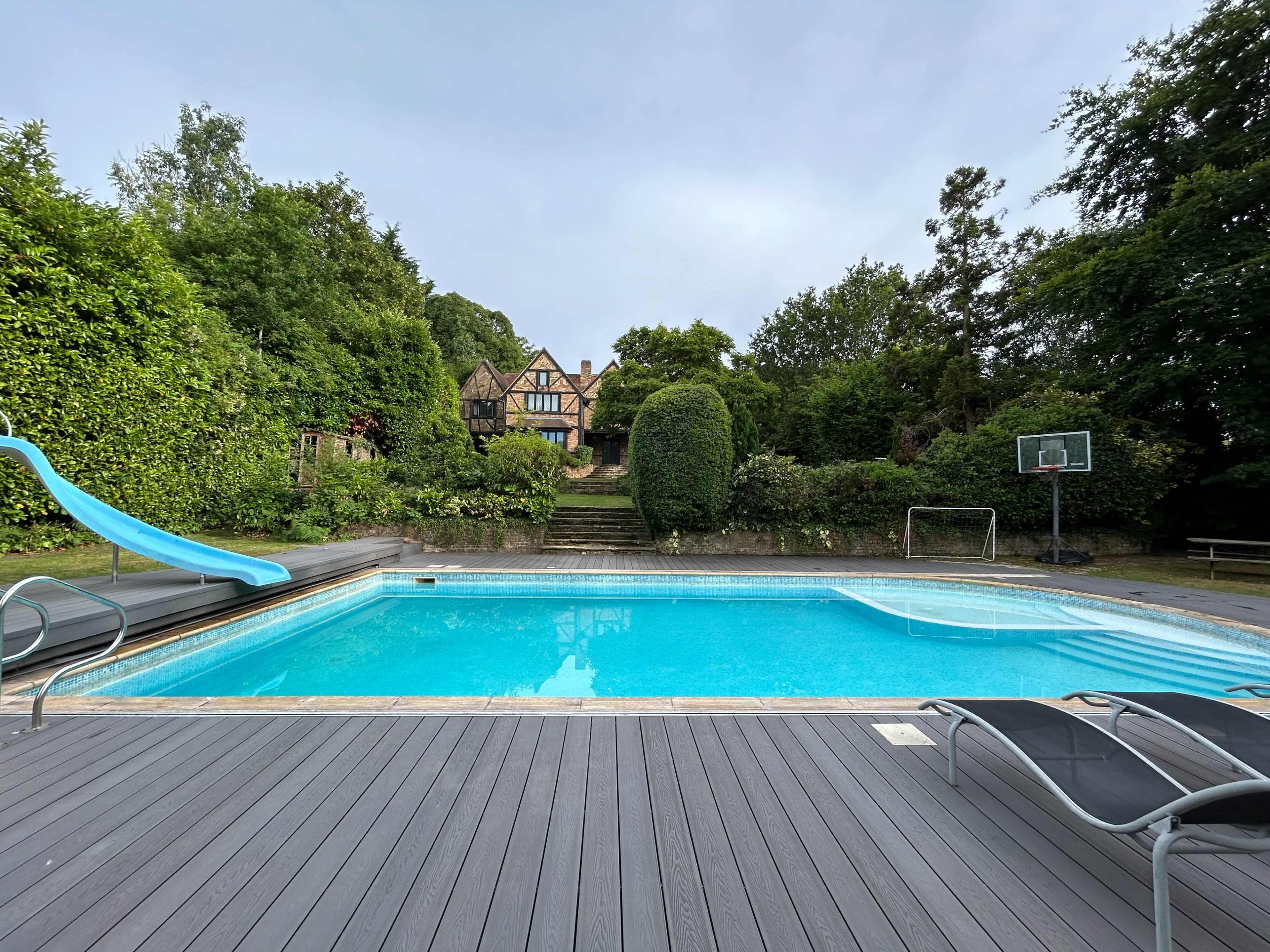 A blue swimming pool with a water slide and lounge chairs is surrounded by lush greenery and a large house in the background.