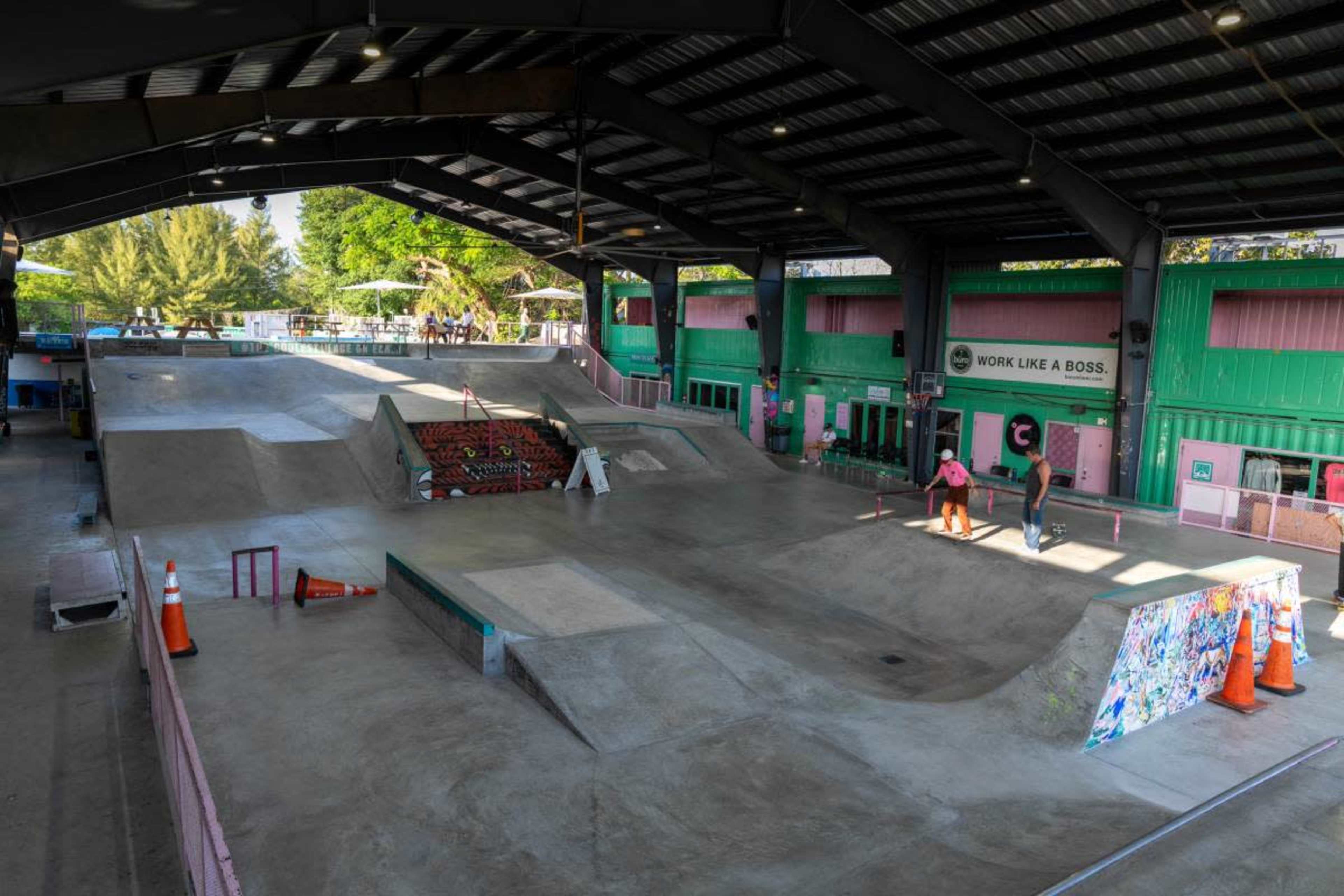 The image shows a large indoor skatepark with concrete ramps, rails, and several skaters in action, surrounded by colorful walls and containers.