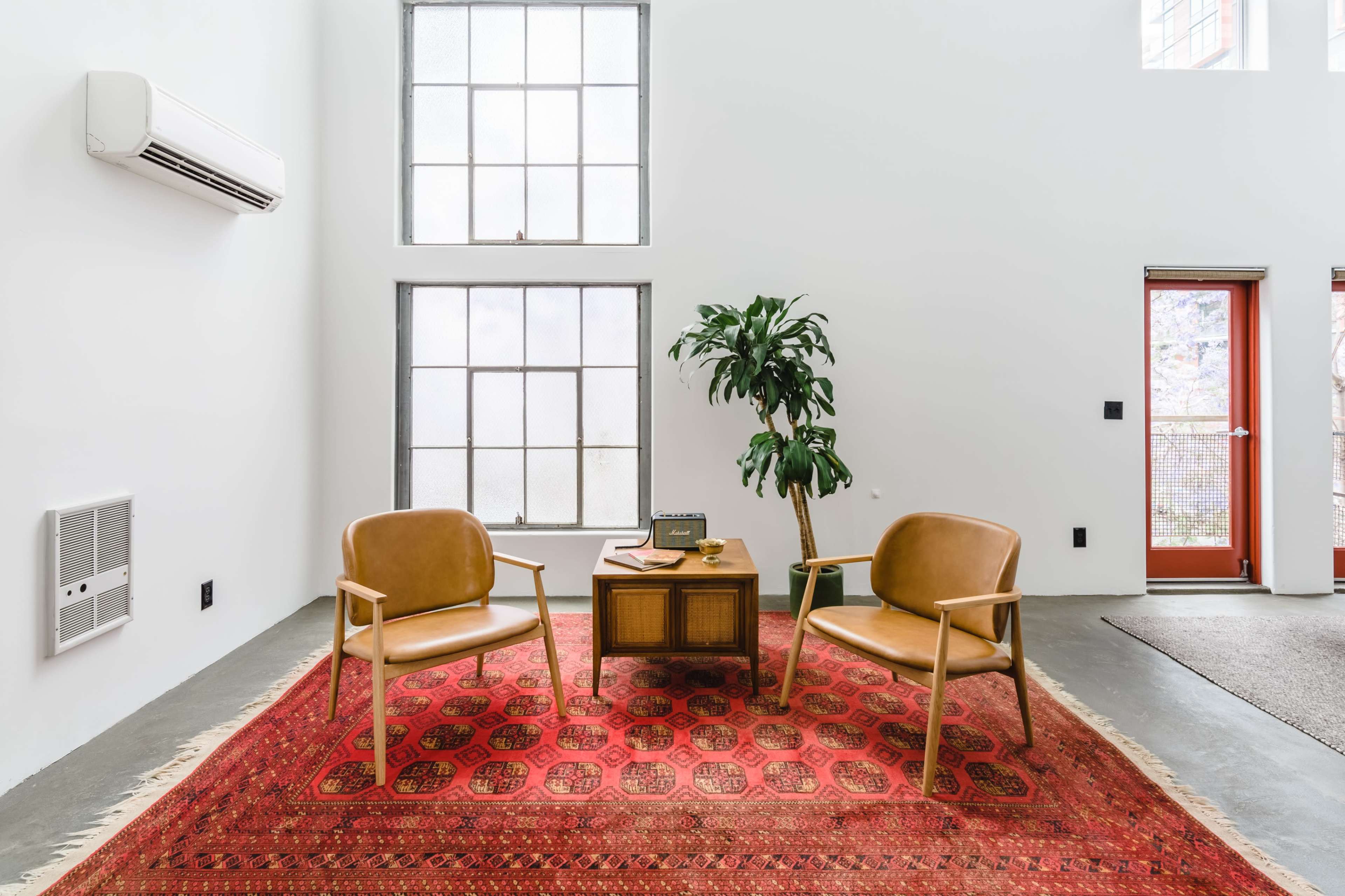 A well-lit interior space features two tan chairs facing a wooden table on a red patterned rug, with large windows and a potted plant nearby.