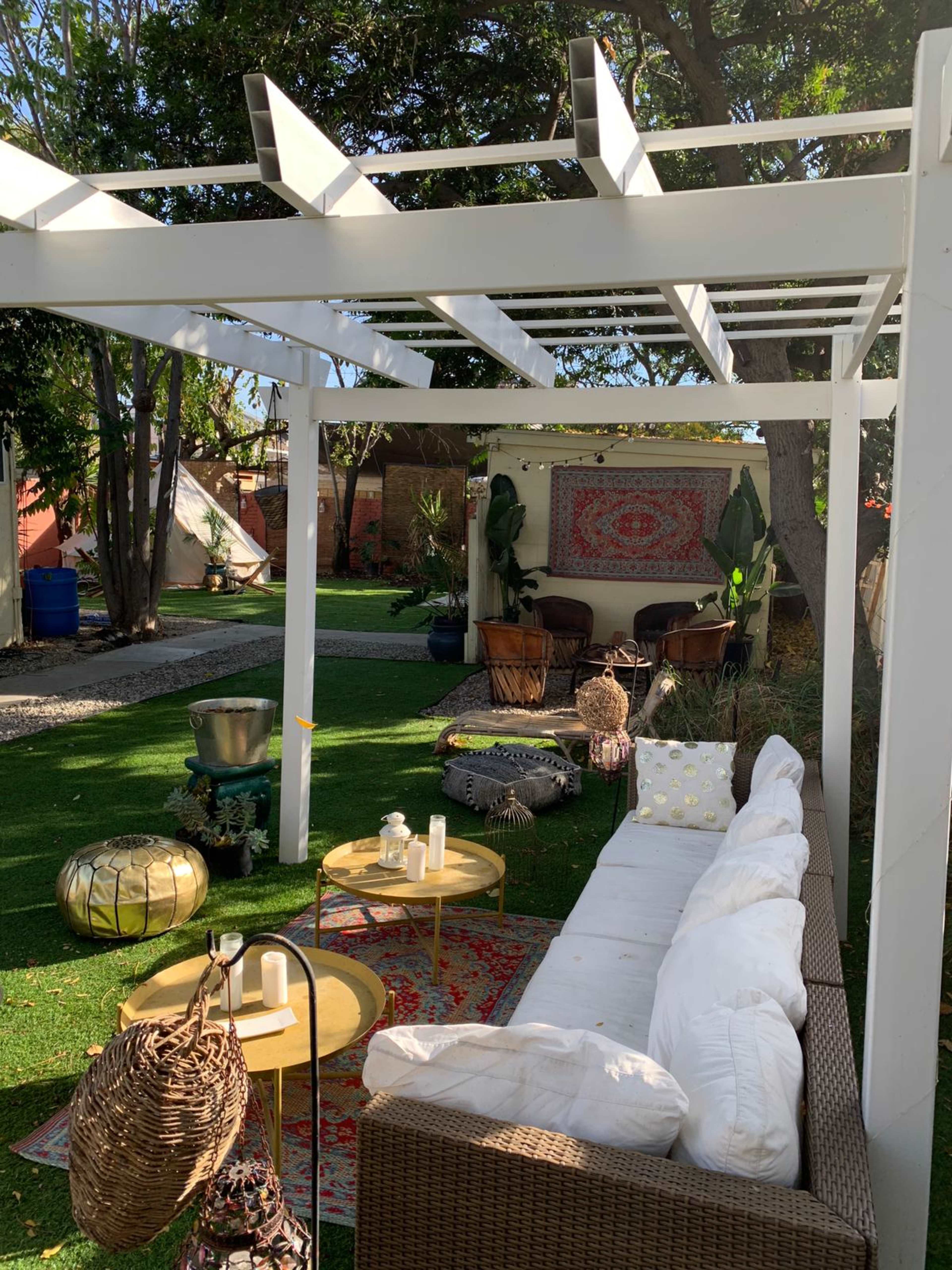 A cozy outdoor seating area under a white pergola, featuring a sofa, decorative tables, and various plants in a landscaped garden.