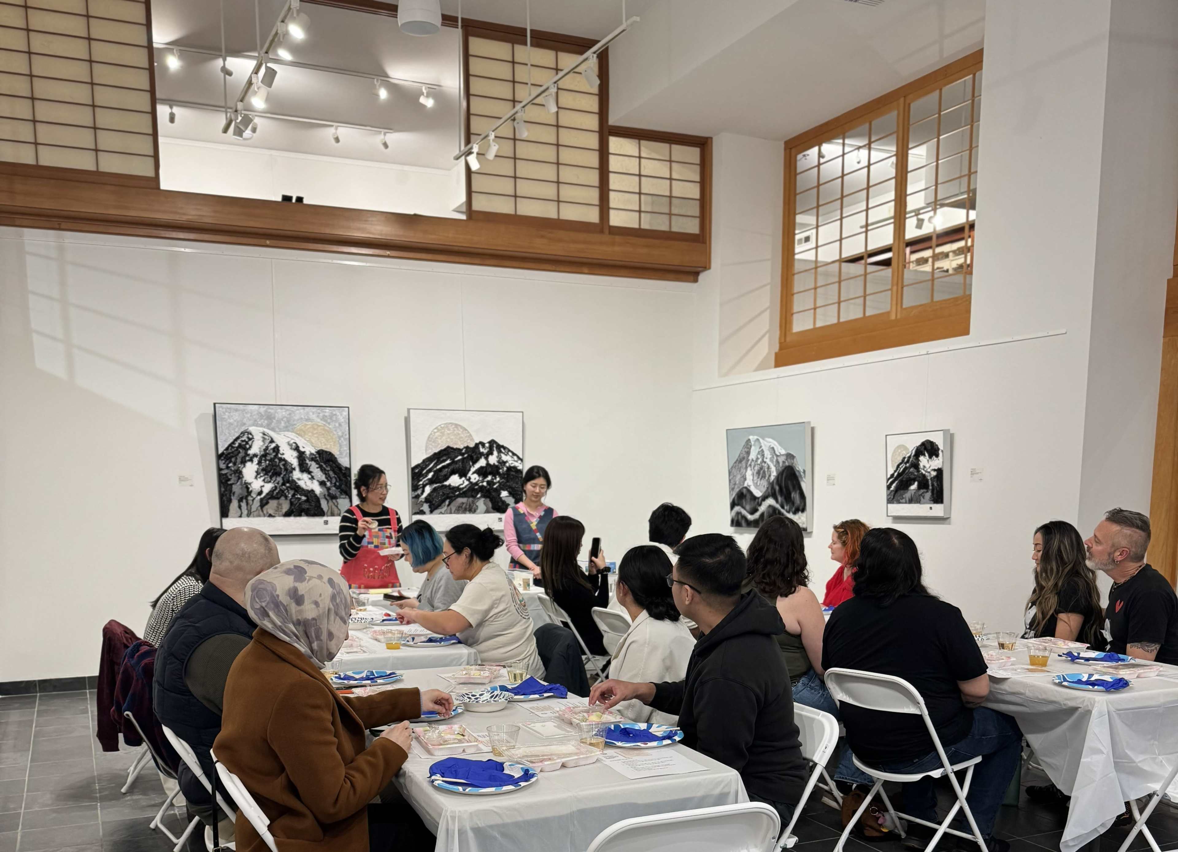A group of people sits at tables in an art gallery, while two individuals speak in front of them, with mountain-themed artworks displayed on the walls.