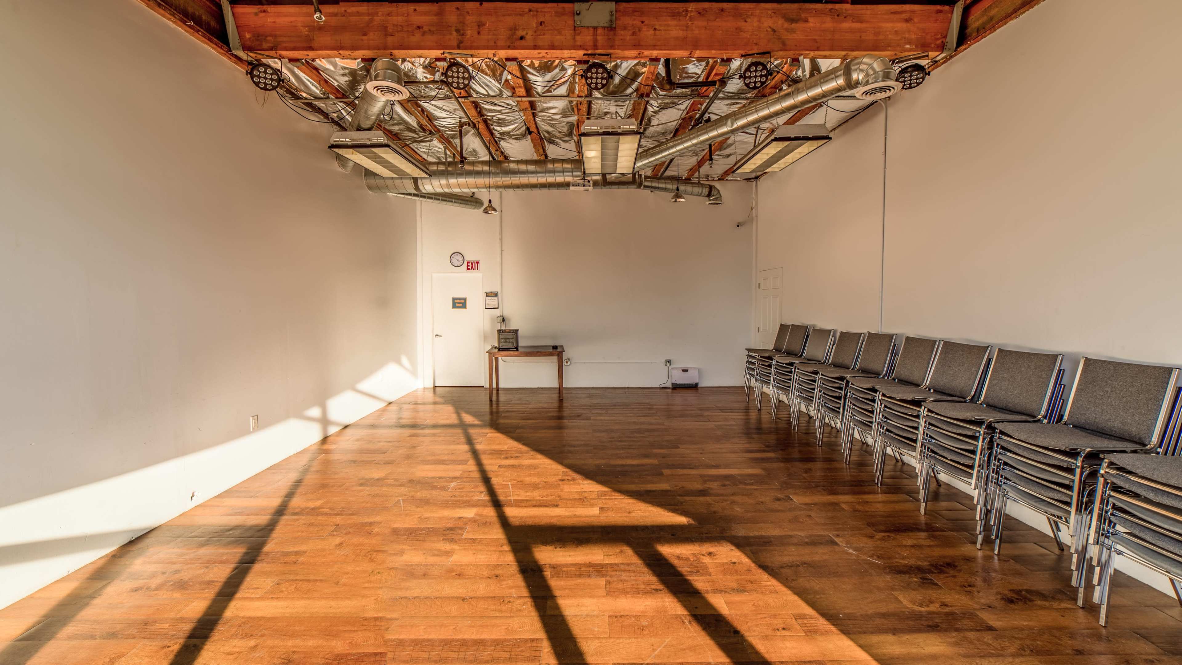 An empty room with hardwood floors, a stack of metal-framed chairs against the wall, and overhead lighting fixtures on a ceiling with exposed beams.