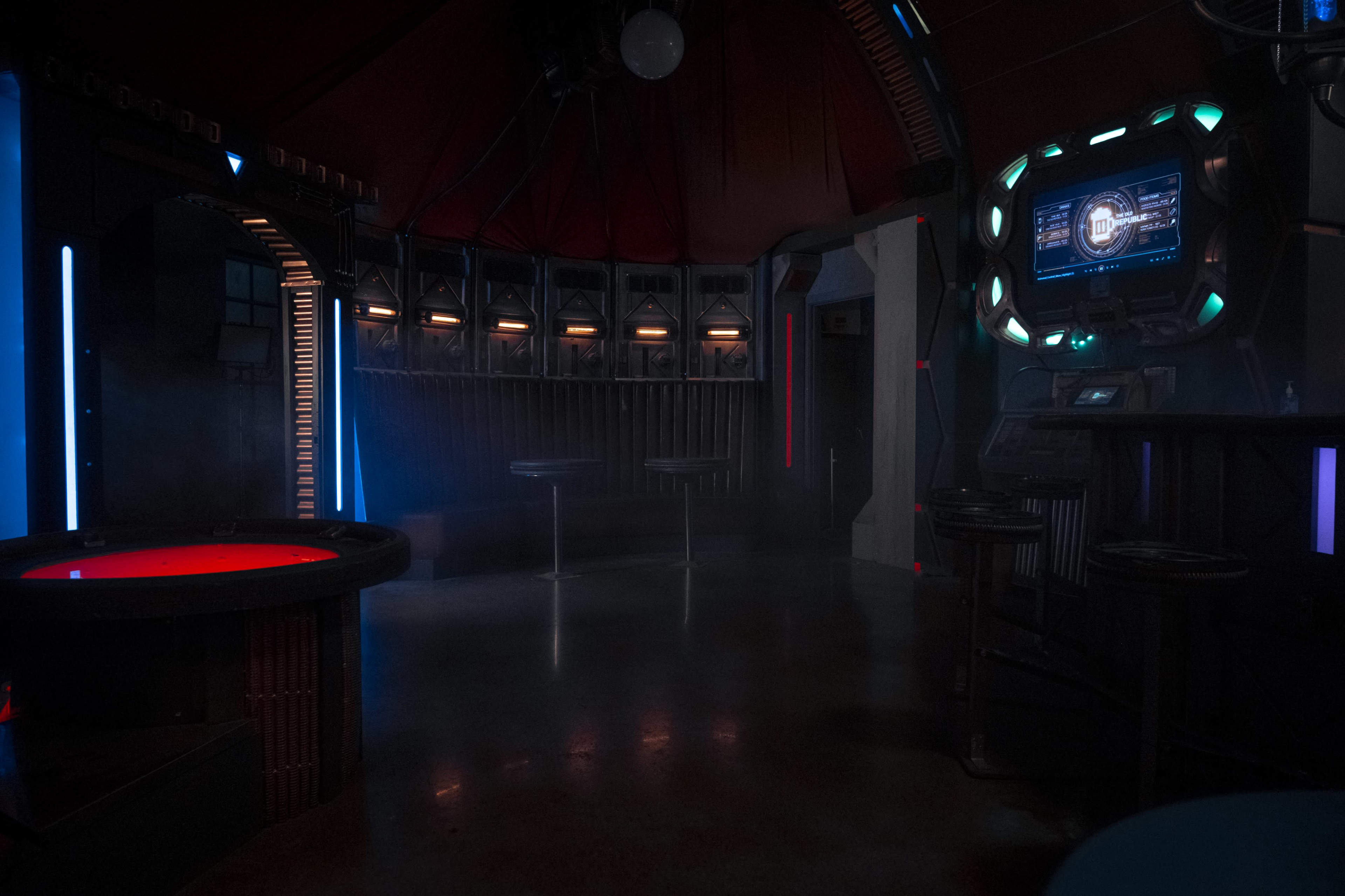 The image depicts a dimly lit futuristic interior with tables, a circular platform emitting red light, and screens displaying digital information on the walls.
