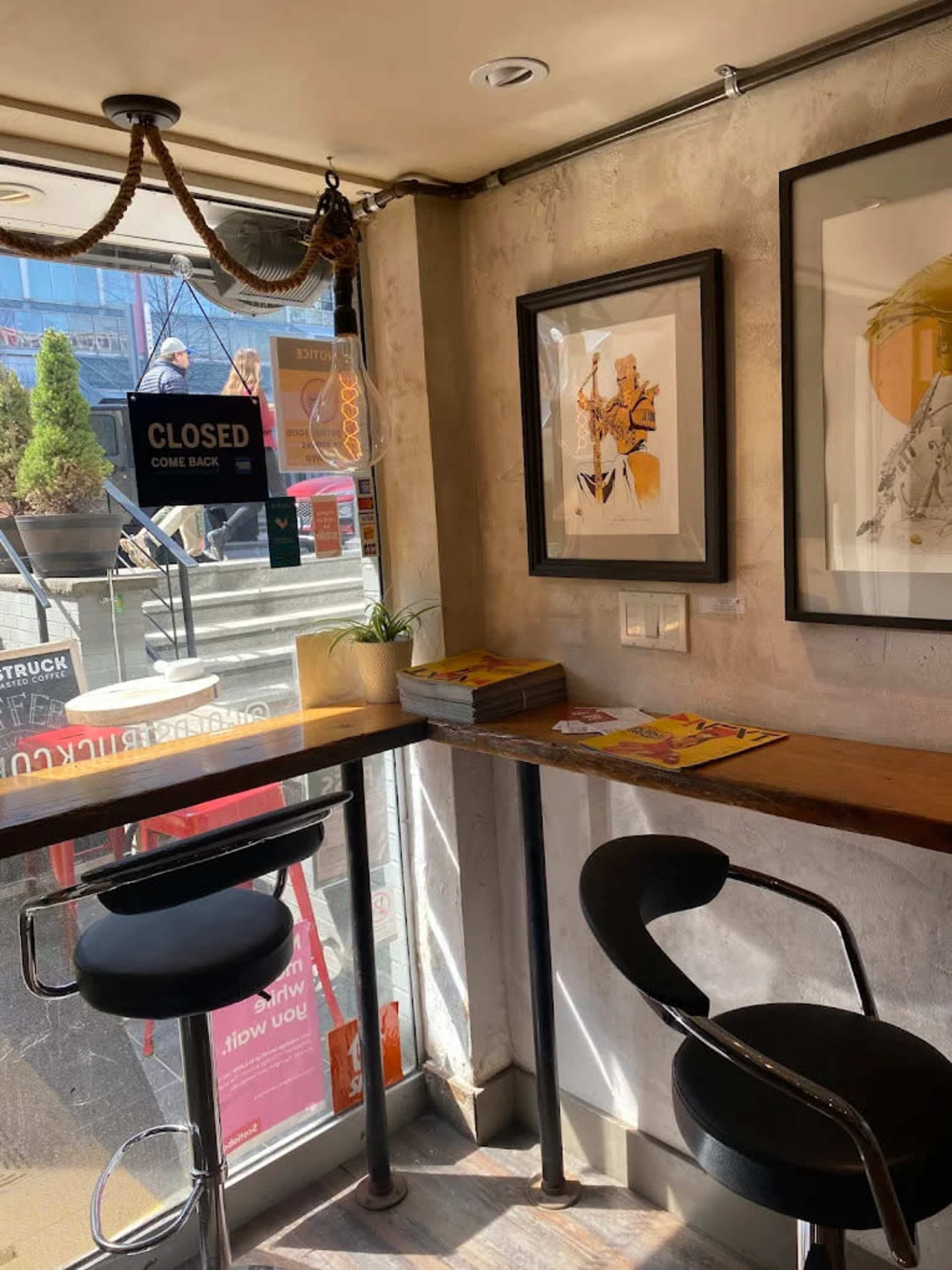The image shows a small café corner with two black chairs and a wooden table, featuring artwork on the wall and a view of steps outside through large windows.