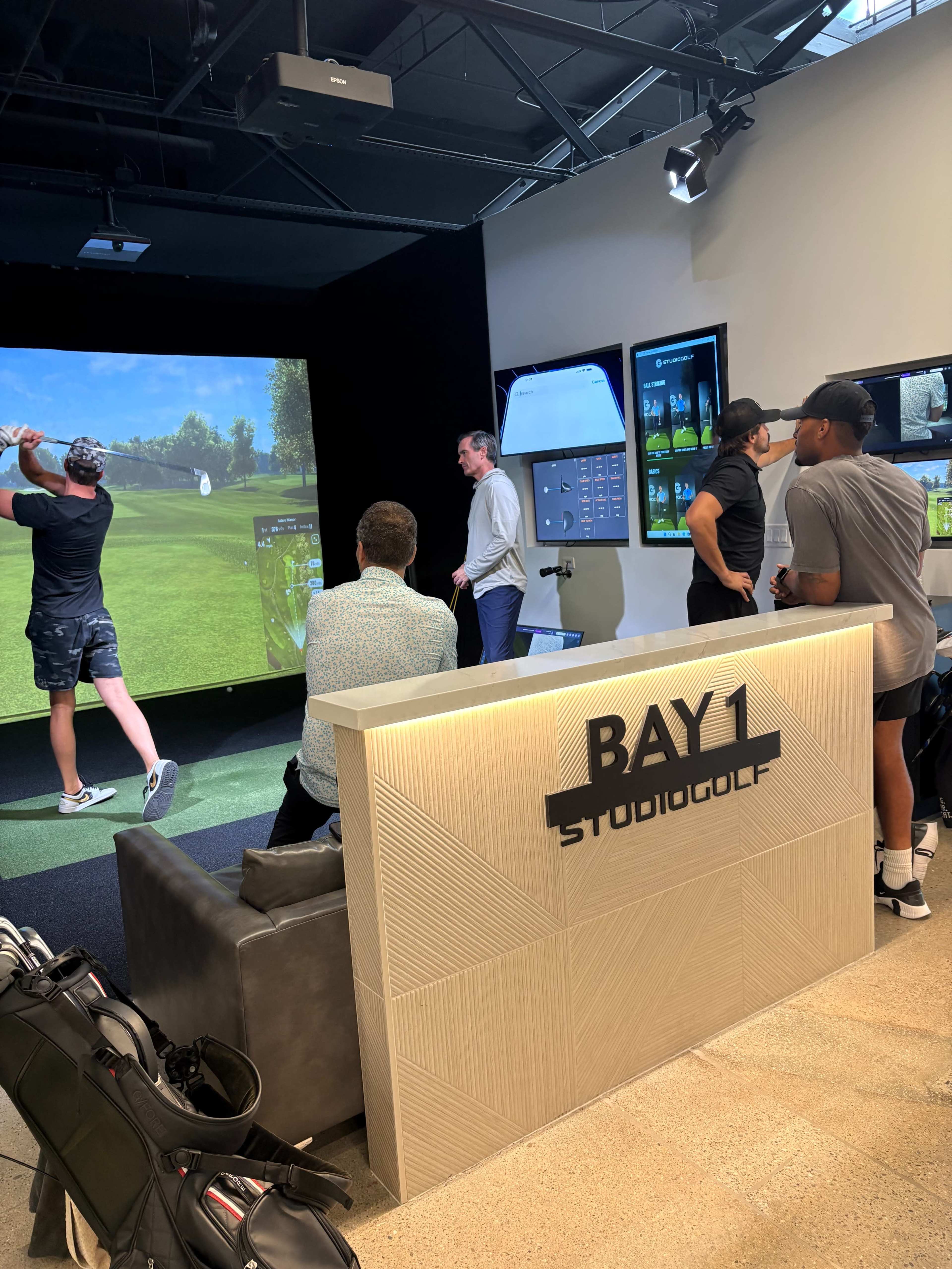 A group of five people observes a golfer using a swing simulator in a golf studio.