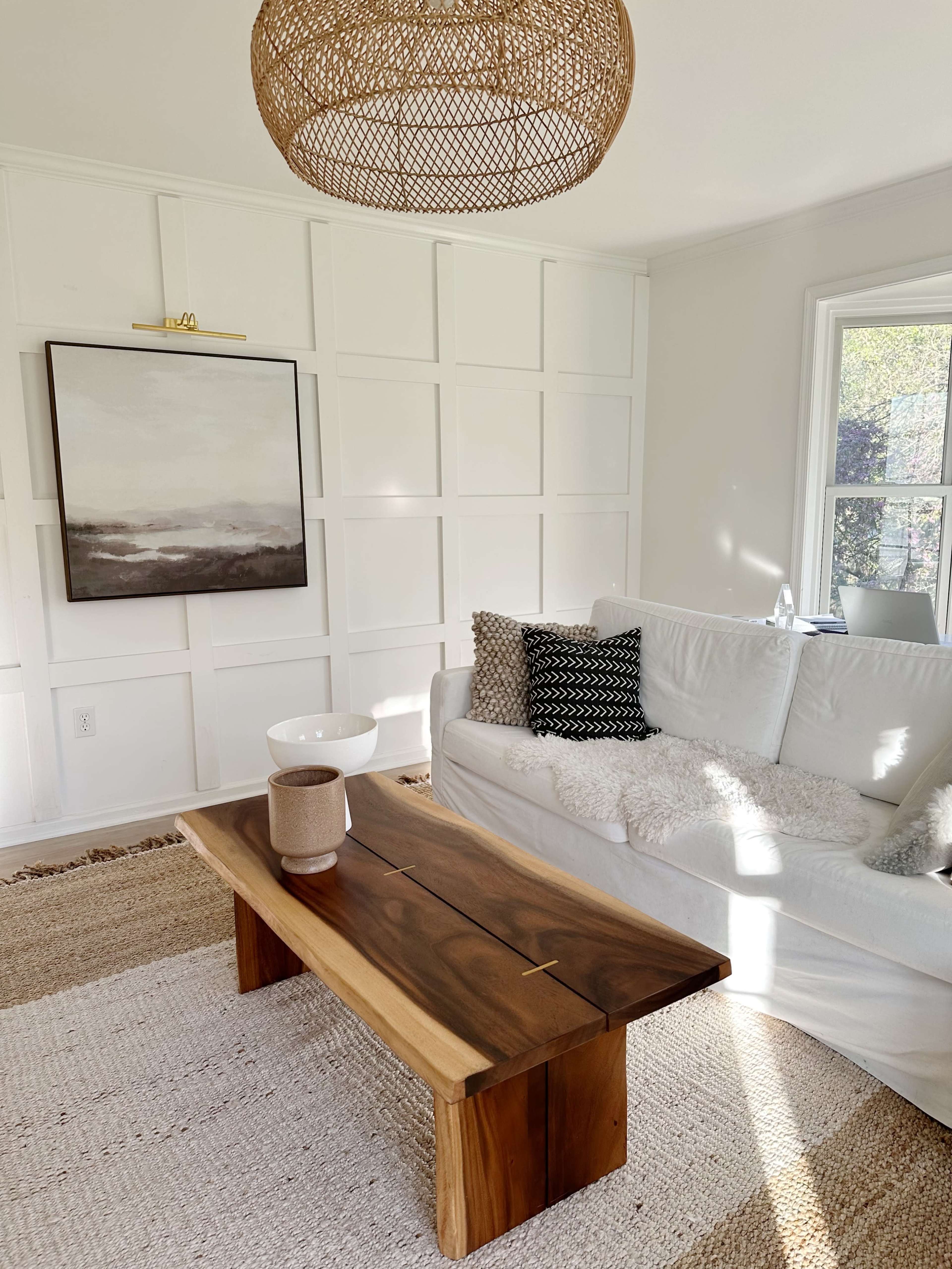 A bright living room features a white sofa adorned with decorative pillows, a wooden coffee table, and a large artwork on the wall, illuminated by a woven pendant light.