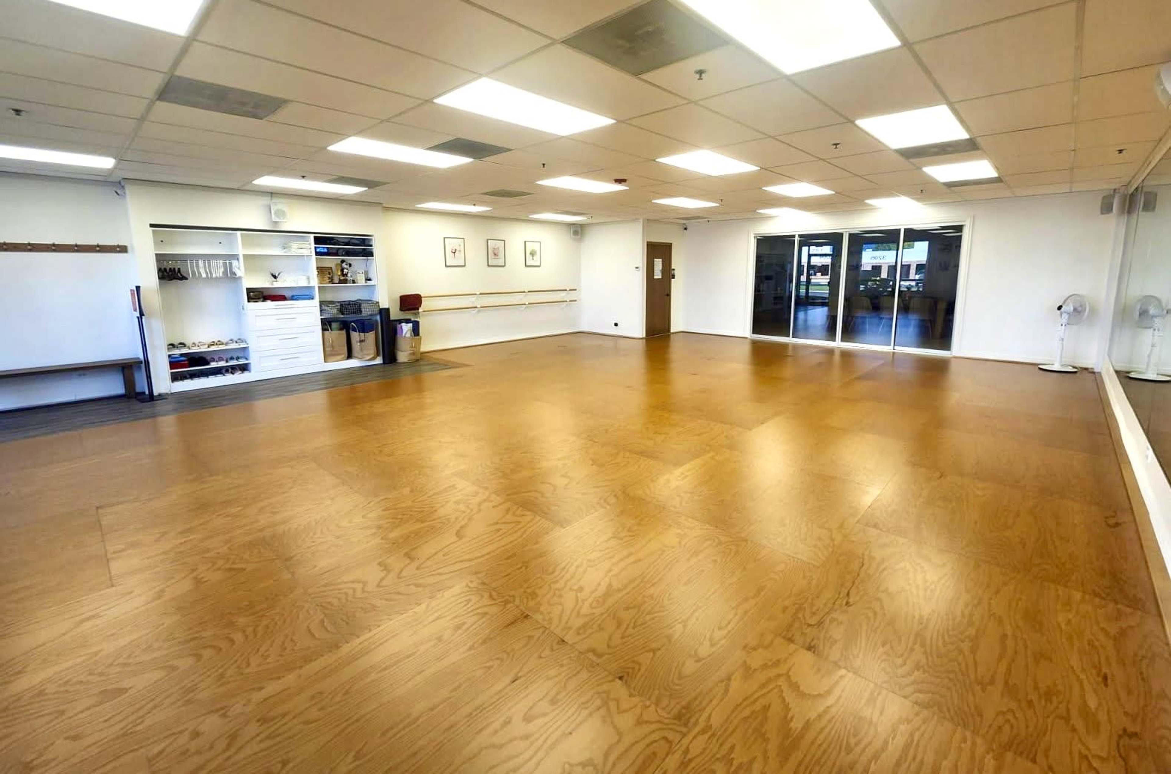 The image shows an empty dance studio with polished wooden floors, large mirrors on one wall, and shelves storing equipment.
