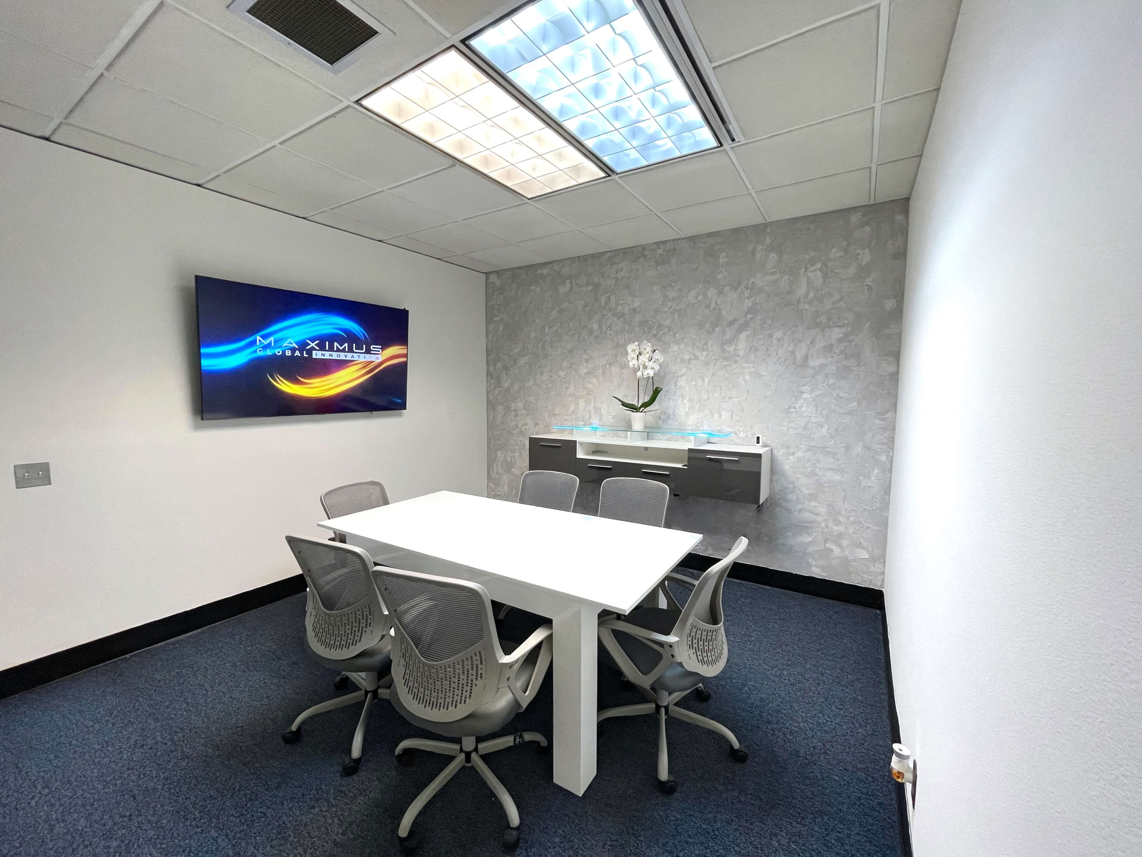 A minimalist conference room features a white table surrounded by gray chairs, with a large screen displaying "MAXIMUS" on one wall.