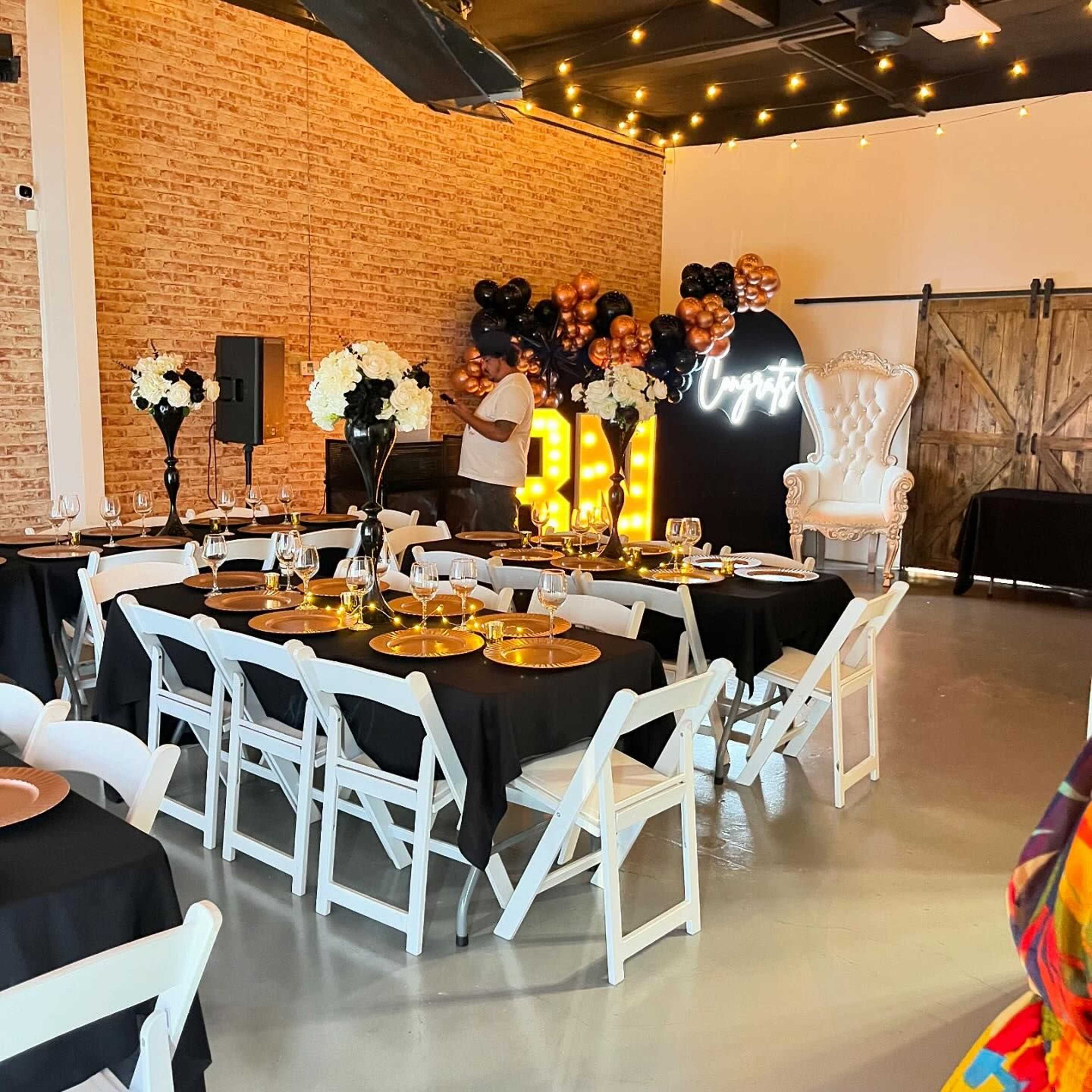 A decorated banquet space with black and white tables, golden accents, and a prominent "50" neon sign amid various balloons.