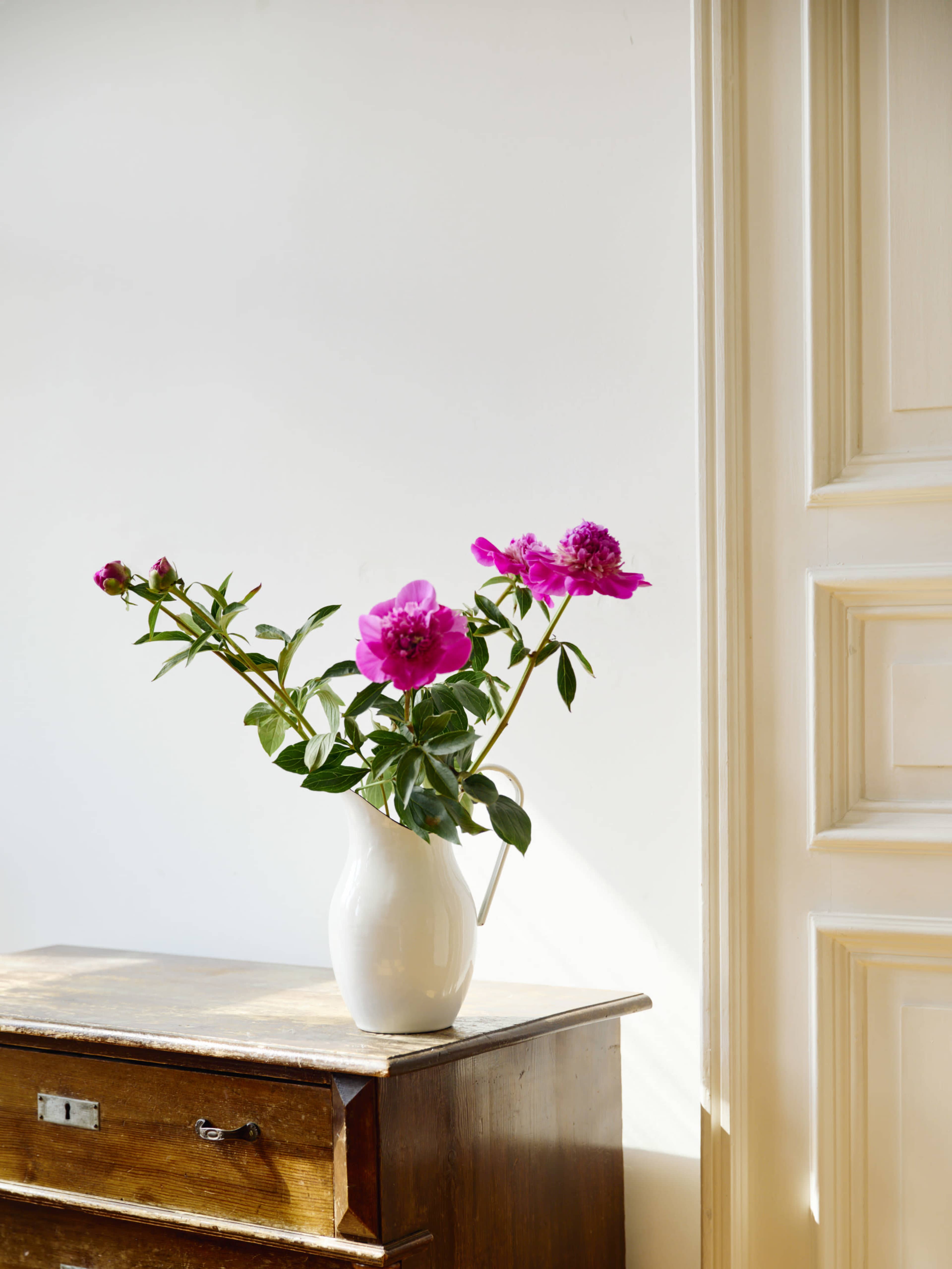 A white vase with pink peonies sits on a wooden table beside a white wall.