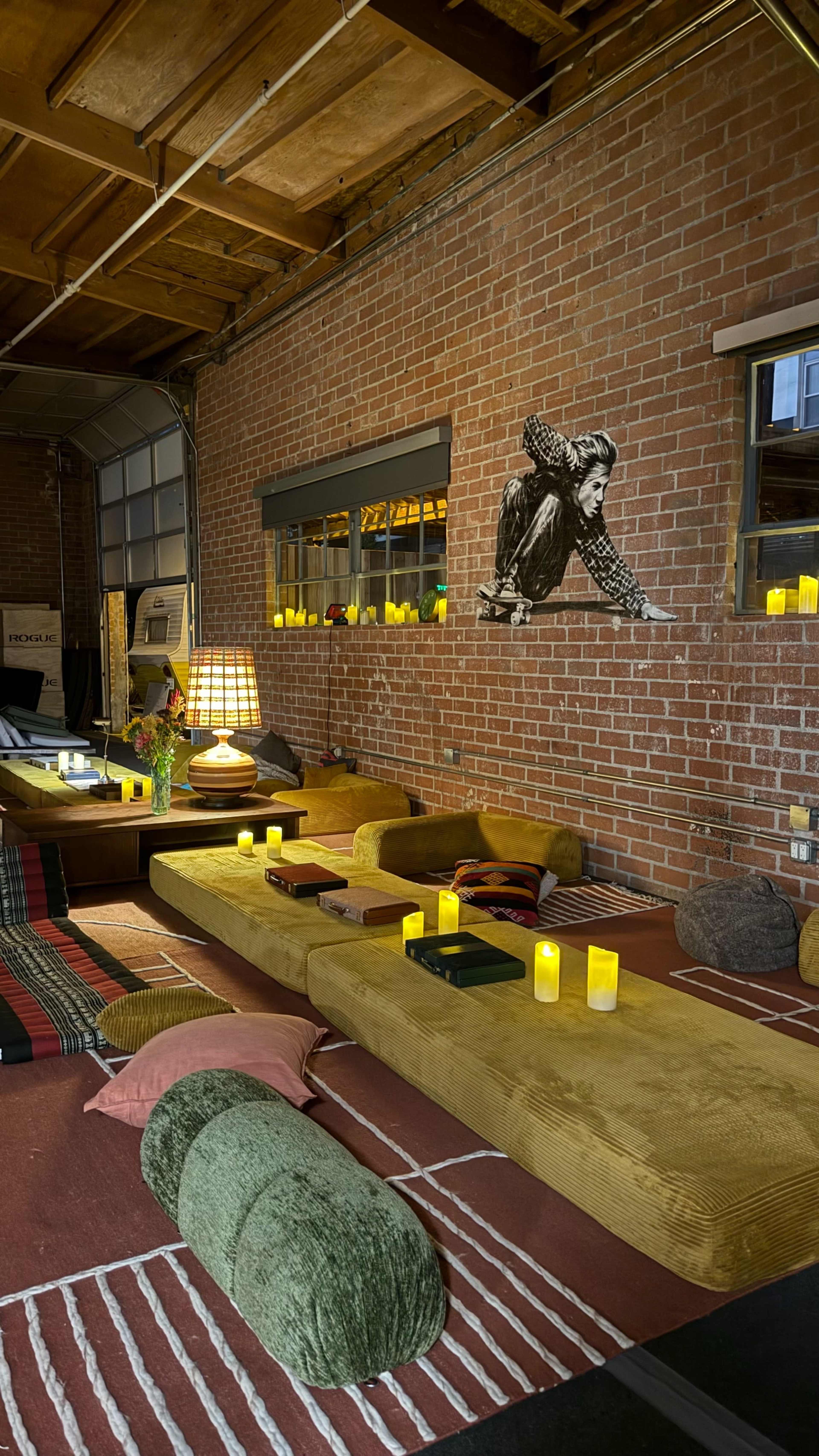 The image shows a cozy, modern interior space with low seating, various cushions, and candles on a table, all complemented by exposed brick walls and ambient lighting.
