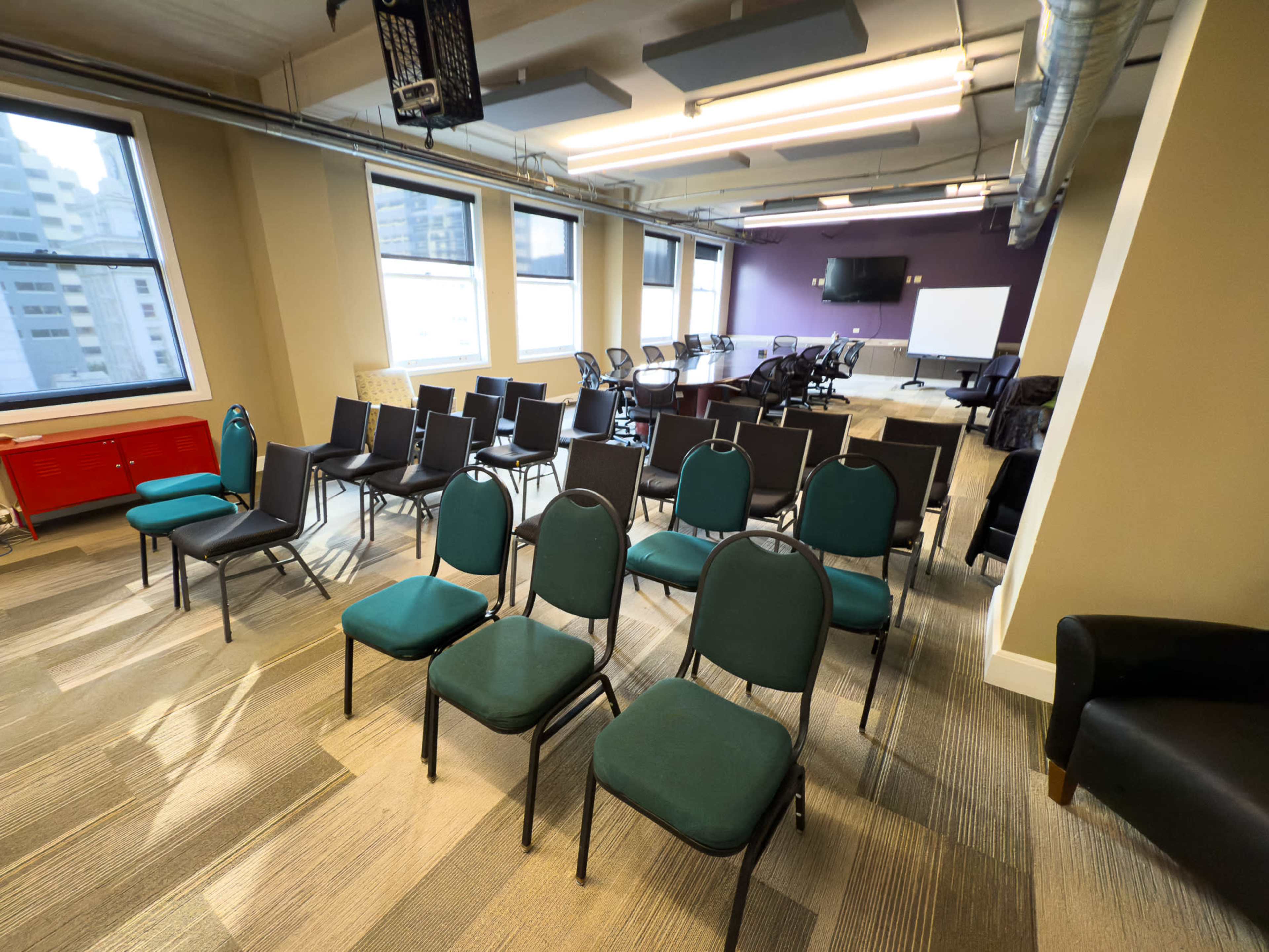 The image shows a conference room with rows of green chairs set up facing a presentation area, accompanied by a few tables and a whiteboard.