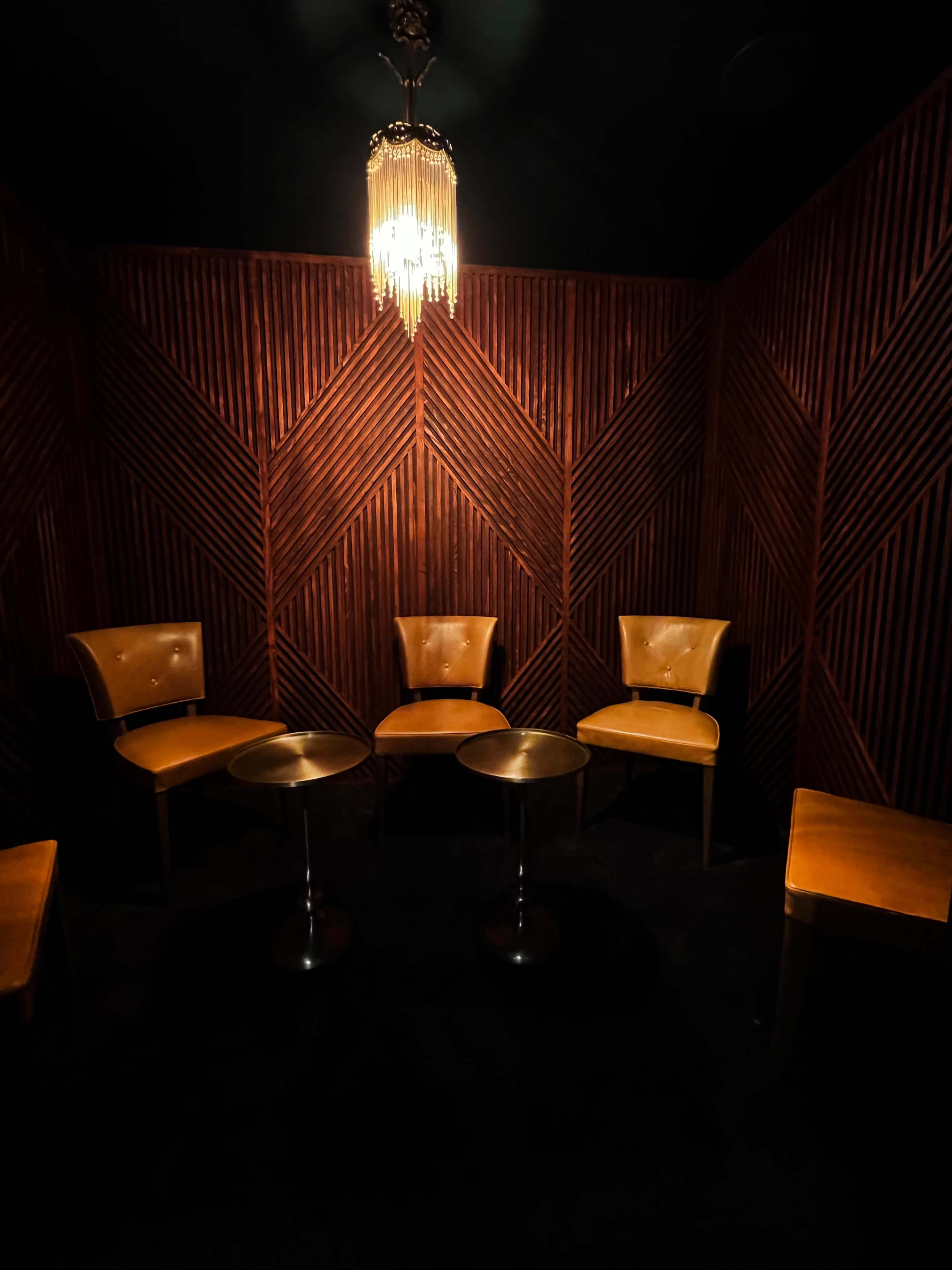 A dimly lit corner of a room with four brown chairs arranged around two small tables, set against wooden paneled walls.