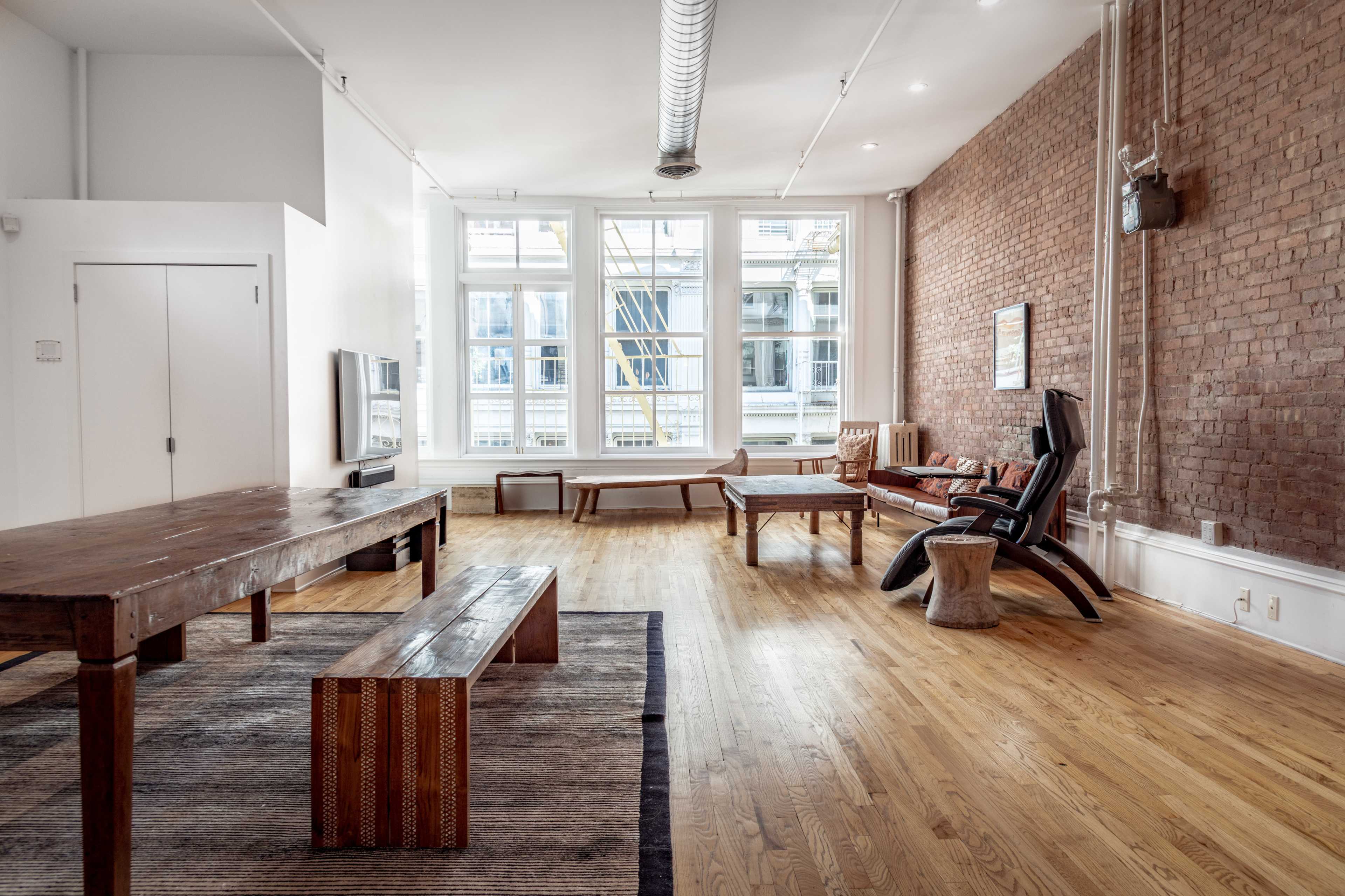 The image shows a spacious room with hardwood flooring, exposed brick walls, a large table, various seating areas, and large windows allowing natural light.