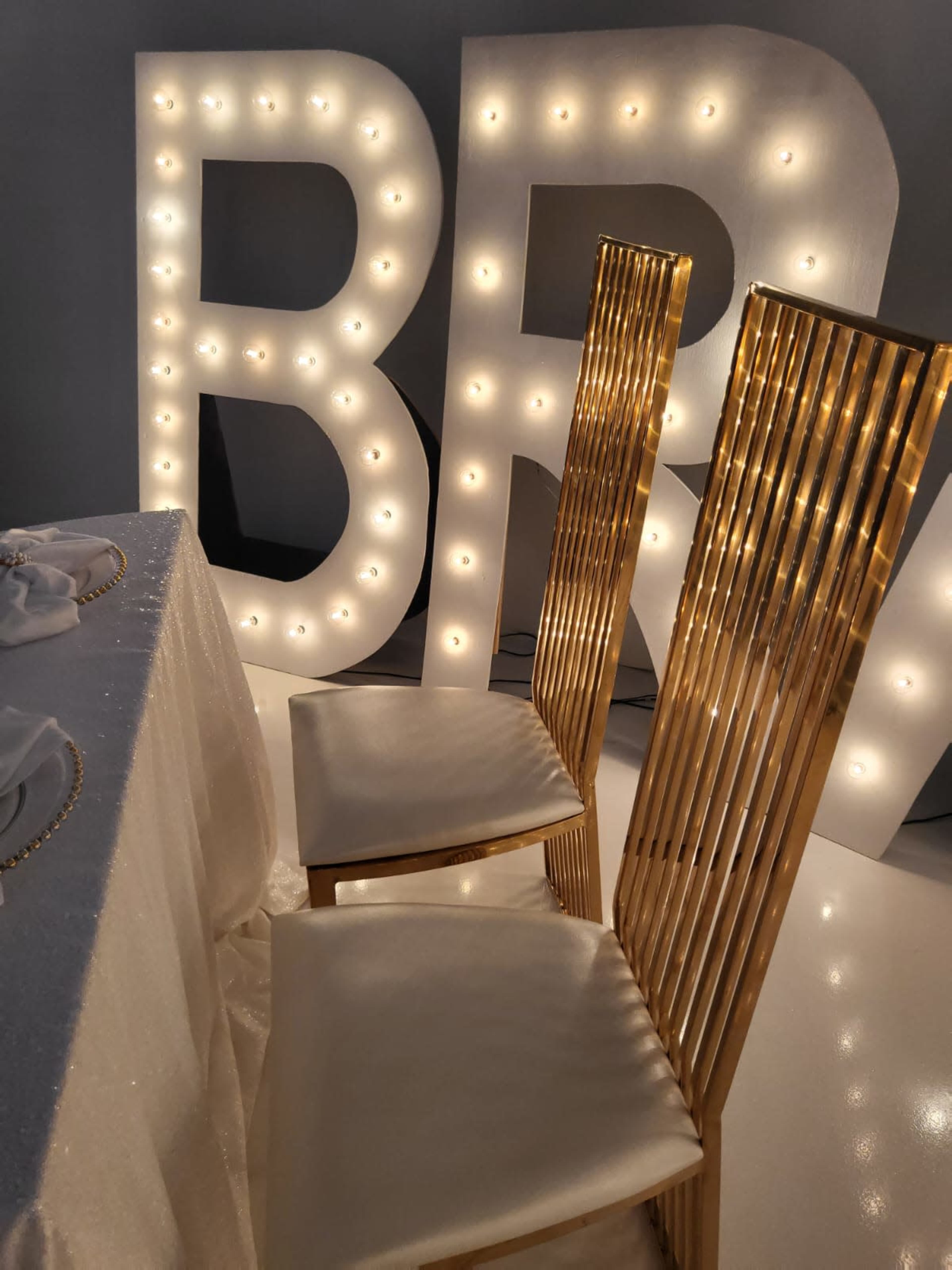 The image shows two elegant gold chairs positioned beside a table, with large illuminated letters spelling "BR" in the background.