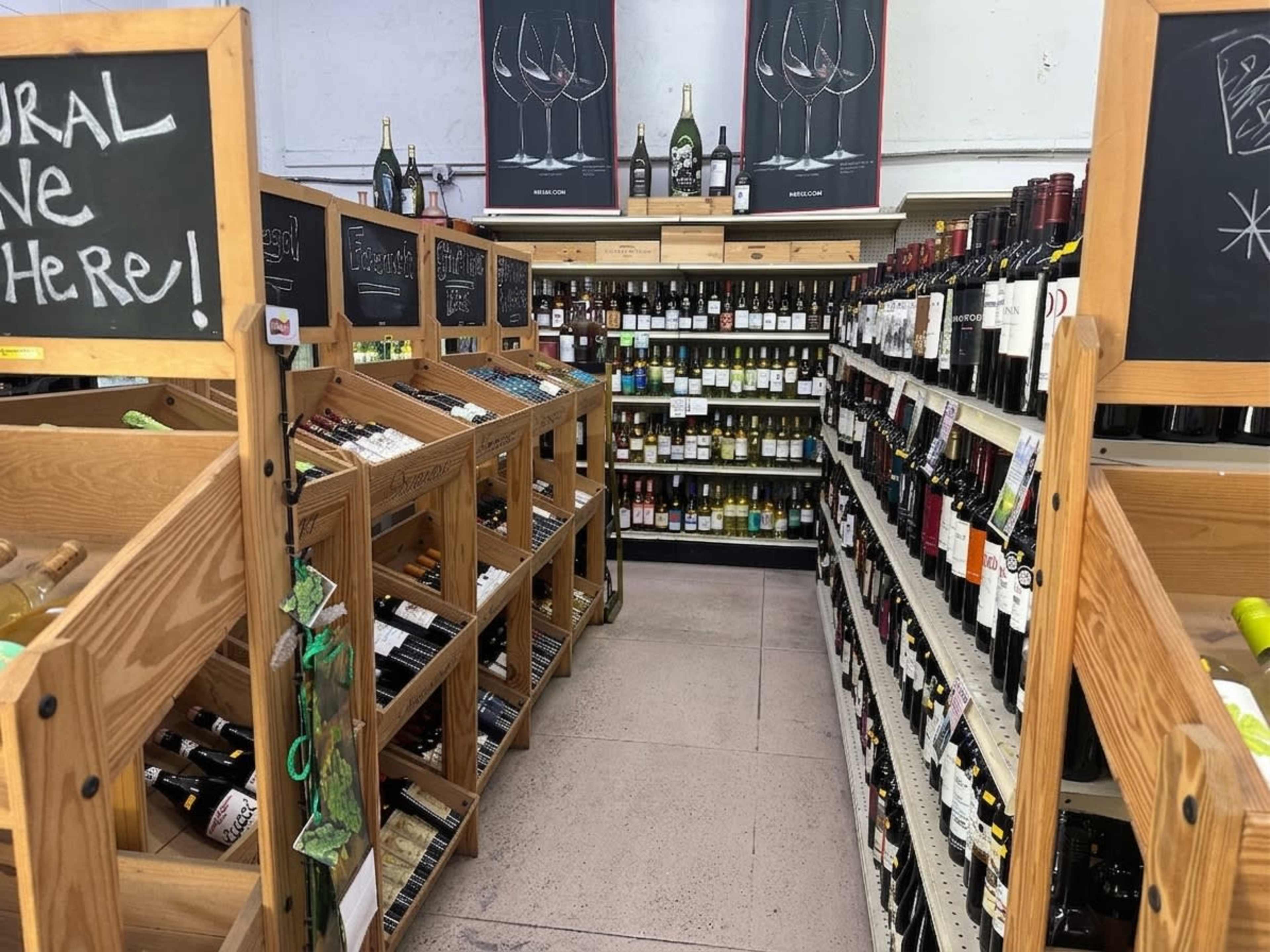 The image shows a wine aisle in a store, featuring wooden display racks filled with various bottles of wine and black chalkboards with text.