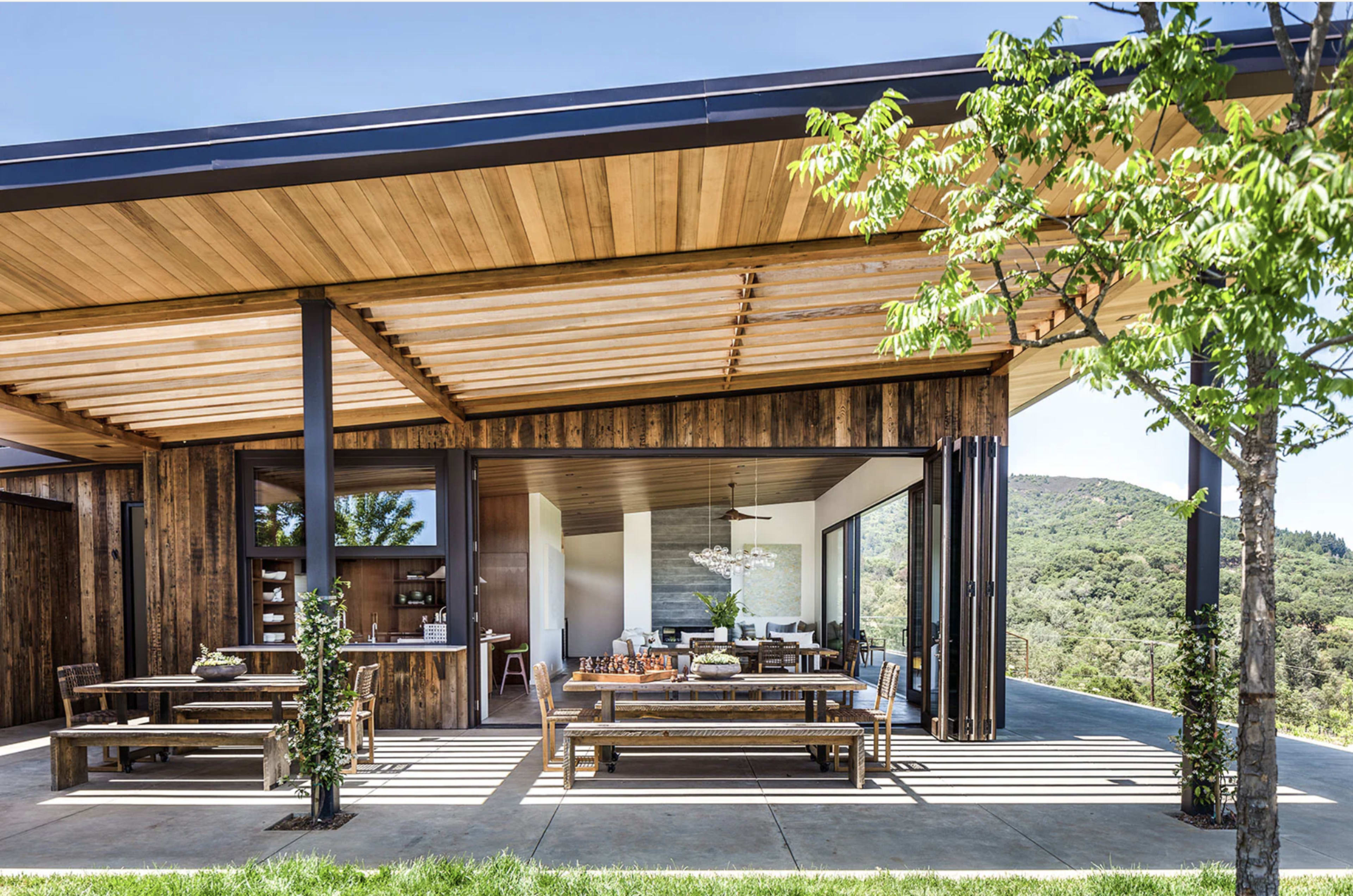 A modern wooden house with large open spaces, featuring outdoor seating and expansive views of greenery.