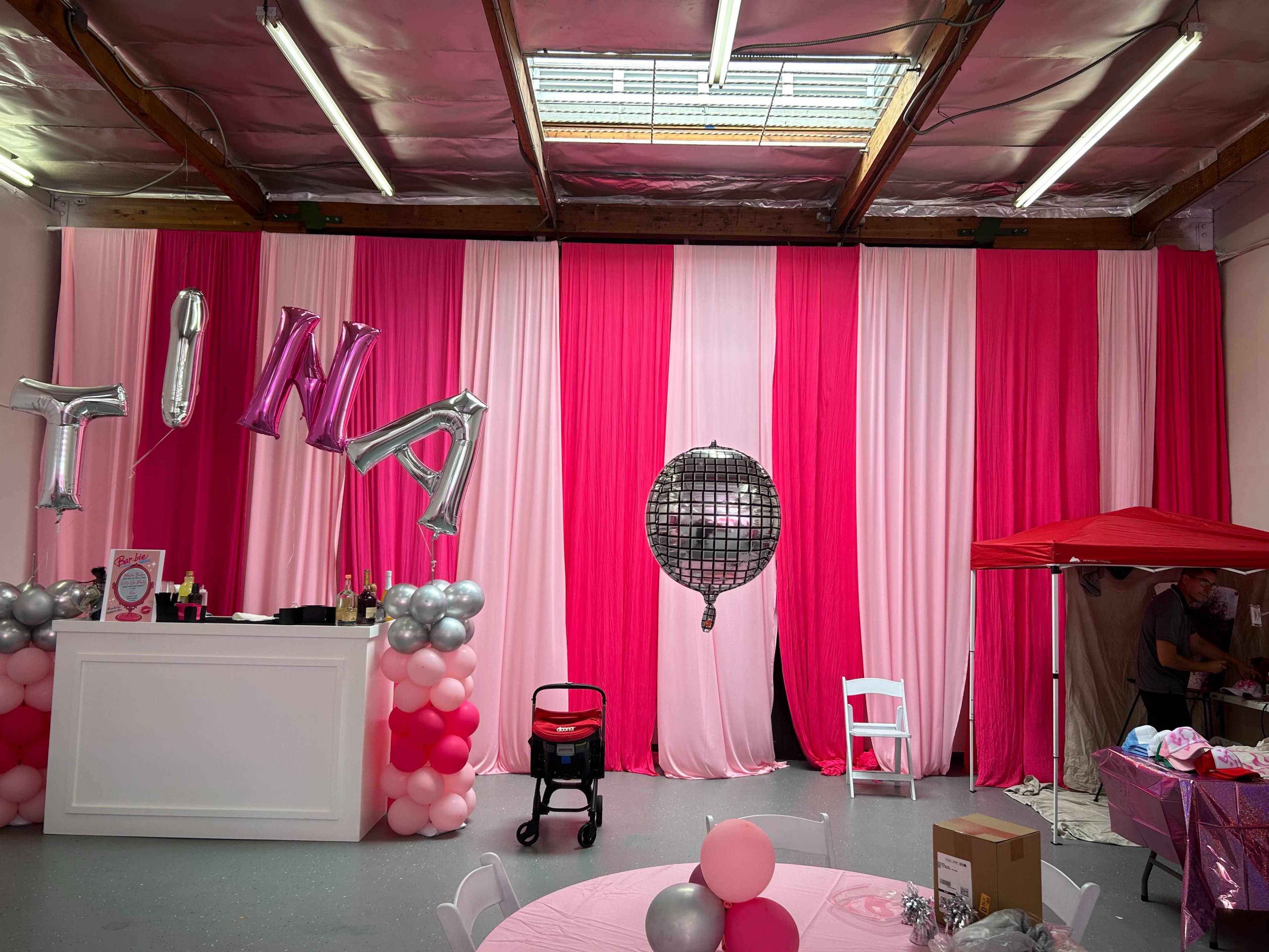 A decorated party space with pink and white curtains, a balloon arch, a bar area, and a disco ball hanging on the wall.
