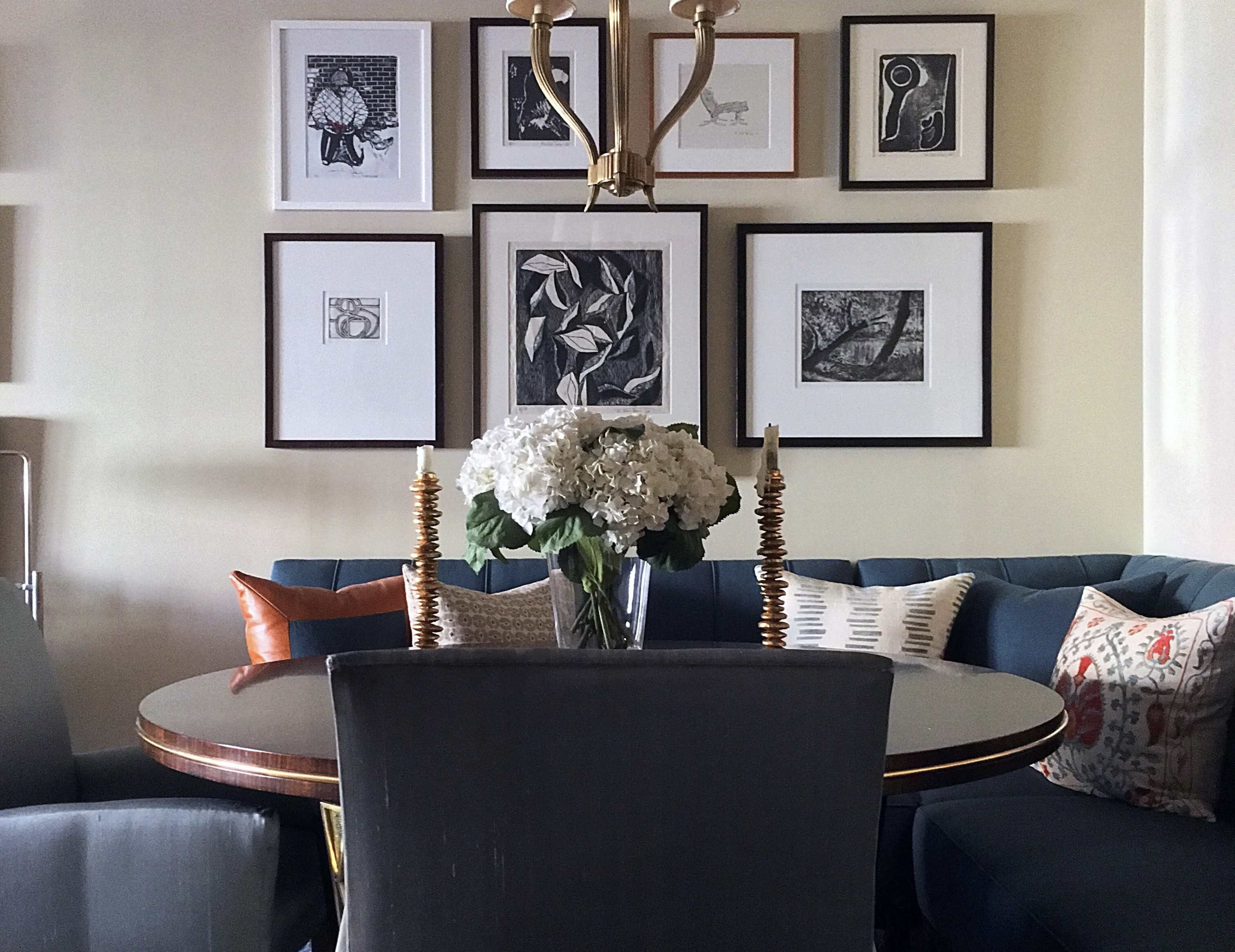 A round dining table with a floral centerpiece is surrounded by upholstered chairs and a gallery wall of framed black-and-white artwork.