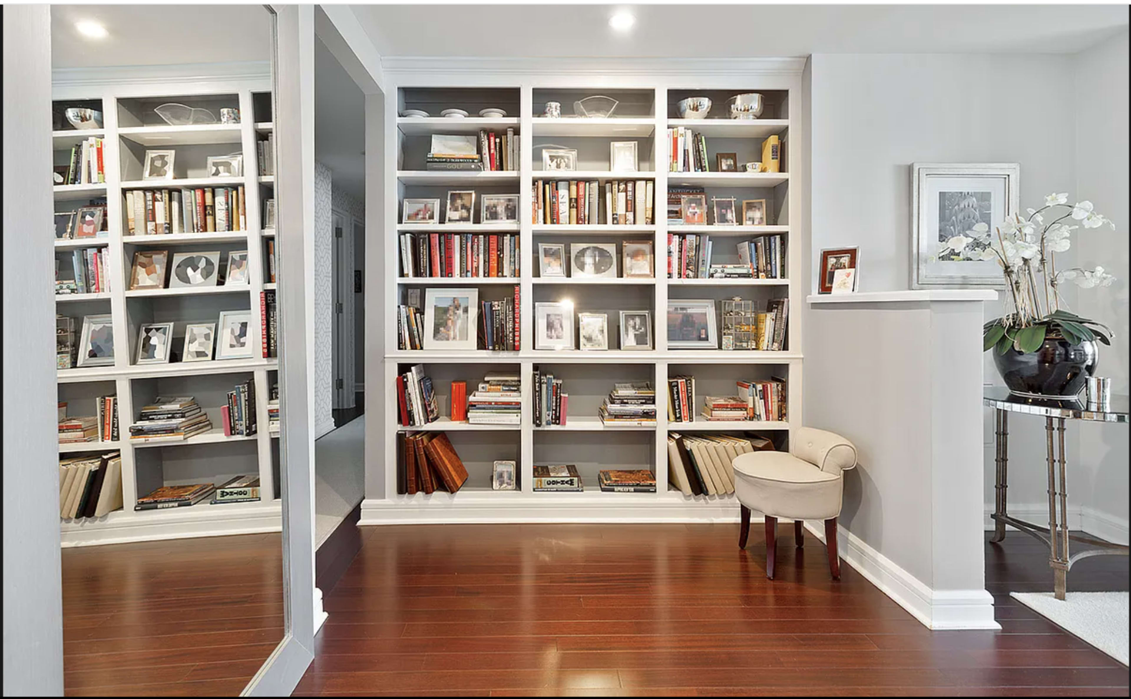 A well-organized bookshelf filled with books and decorative items, alongside a small chair and a mirror reflecting the space.