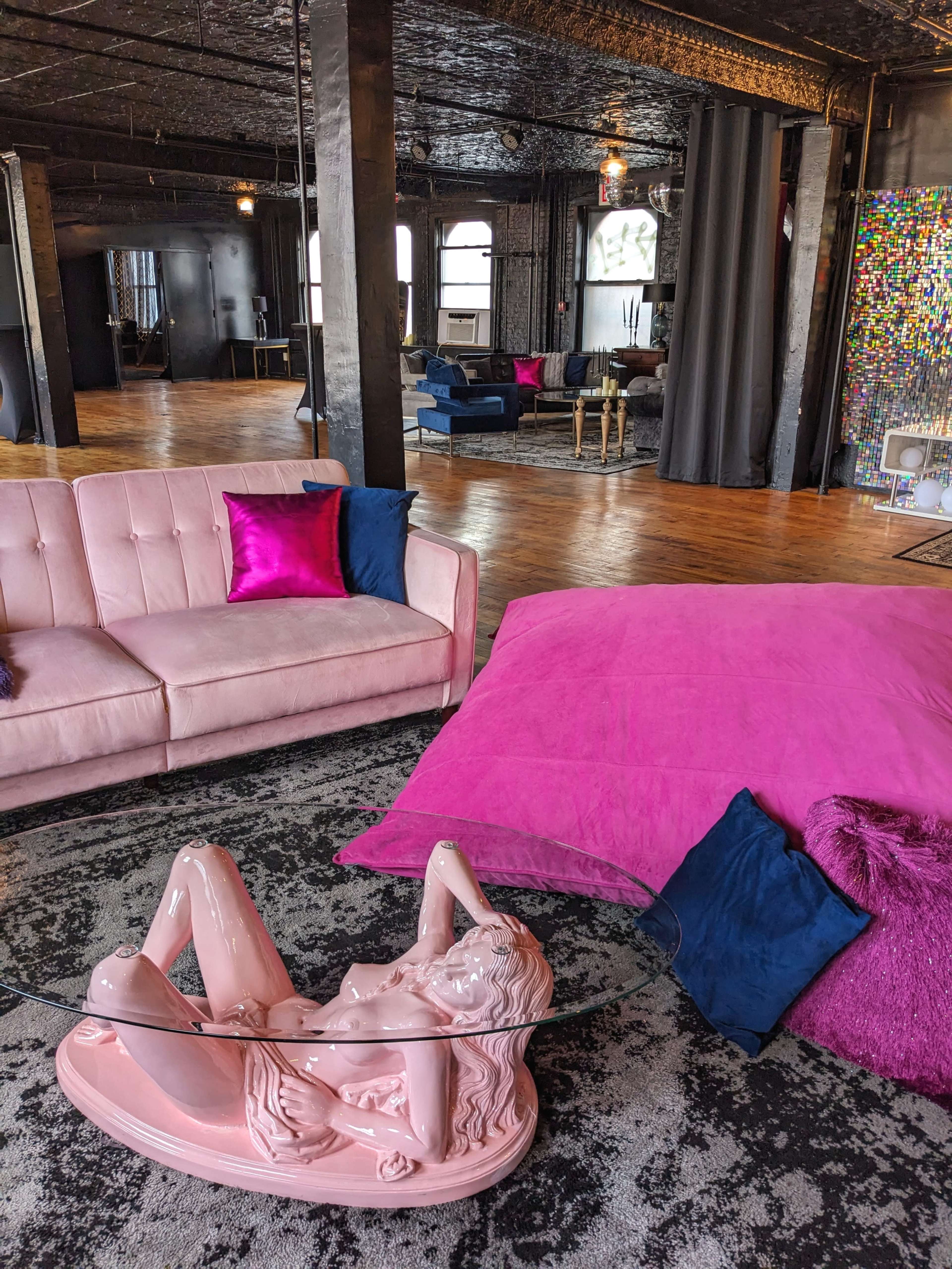 A modern interior space featuring a pink couch, a large pink cushion, and a unique glass table with a sculpture underneath, all against a backdrop of dark walls and wooden flooring.