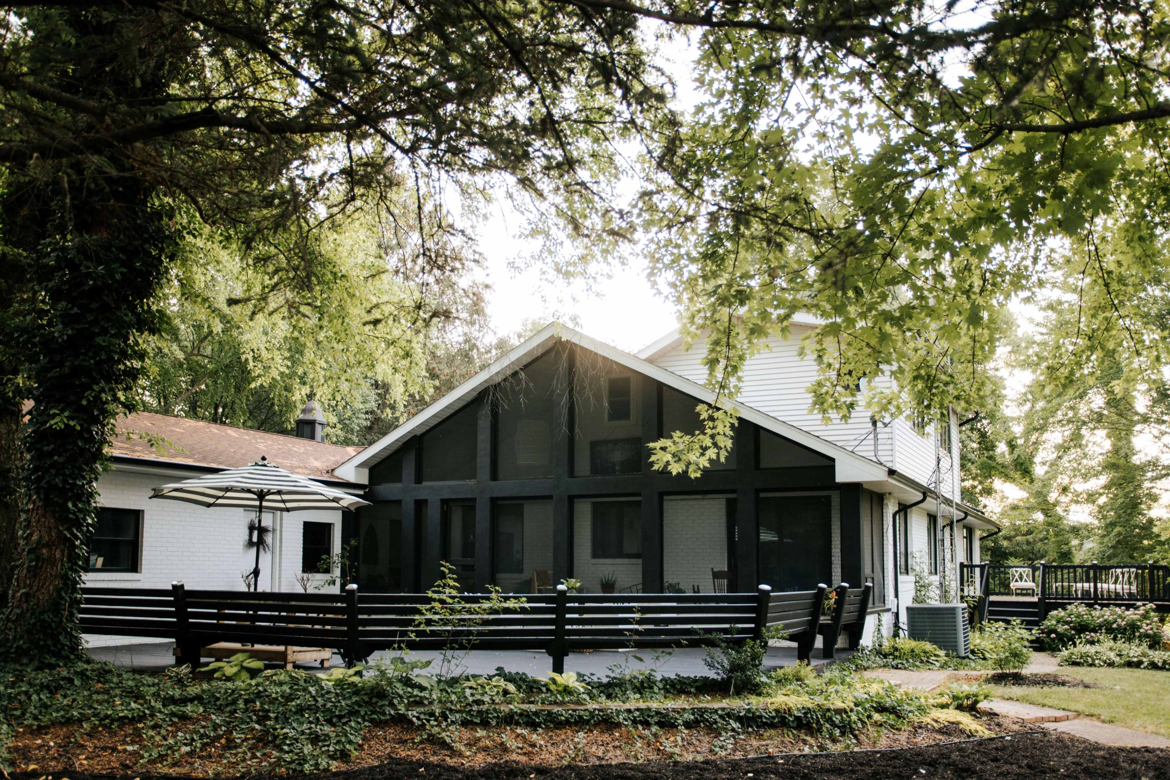 A two-story house with a large front porch is surrounded by trees and landscaping, featuring a deck and an umbrella table in the yard.