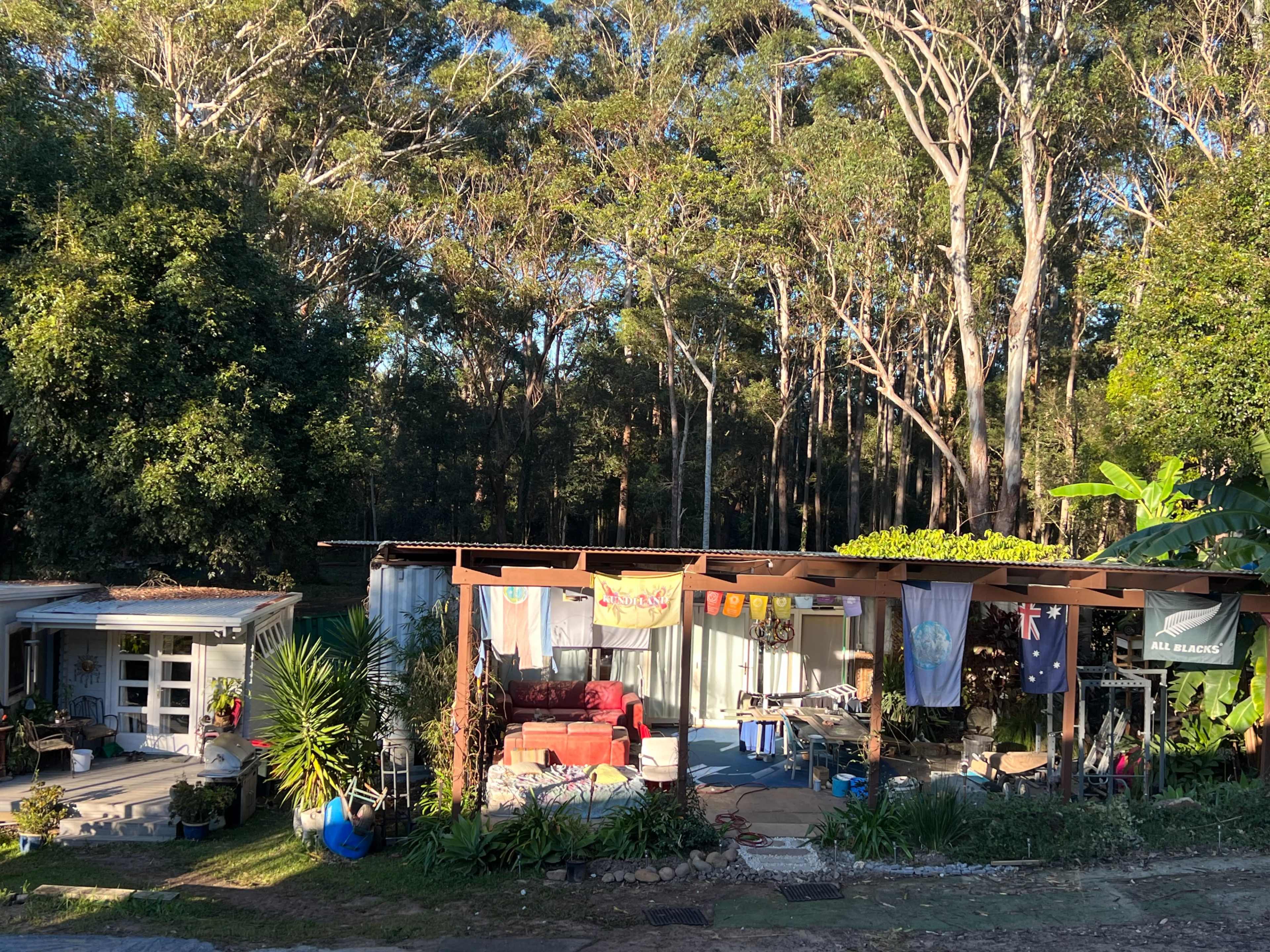 A rustic outdoor living space with various flags, plants, and furniture, situated in a forested area.