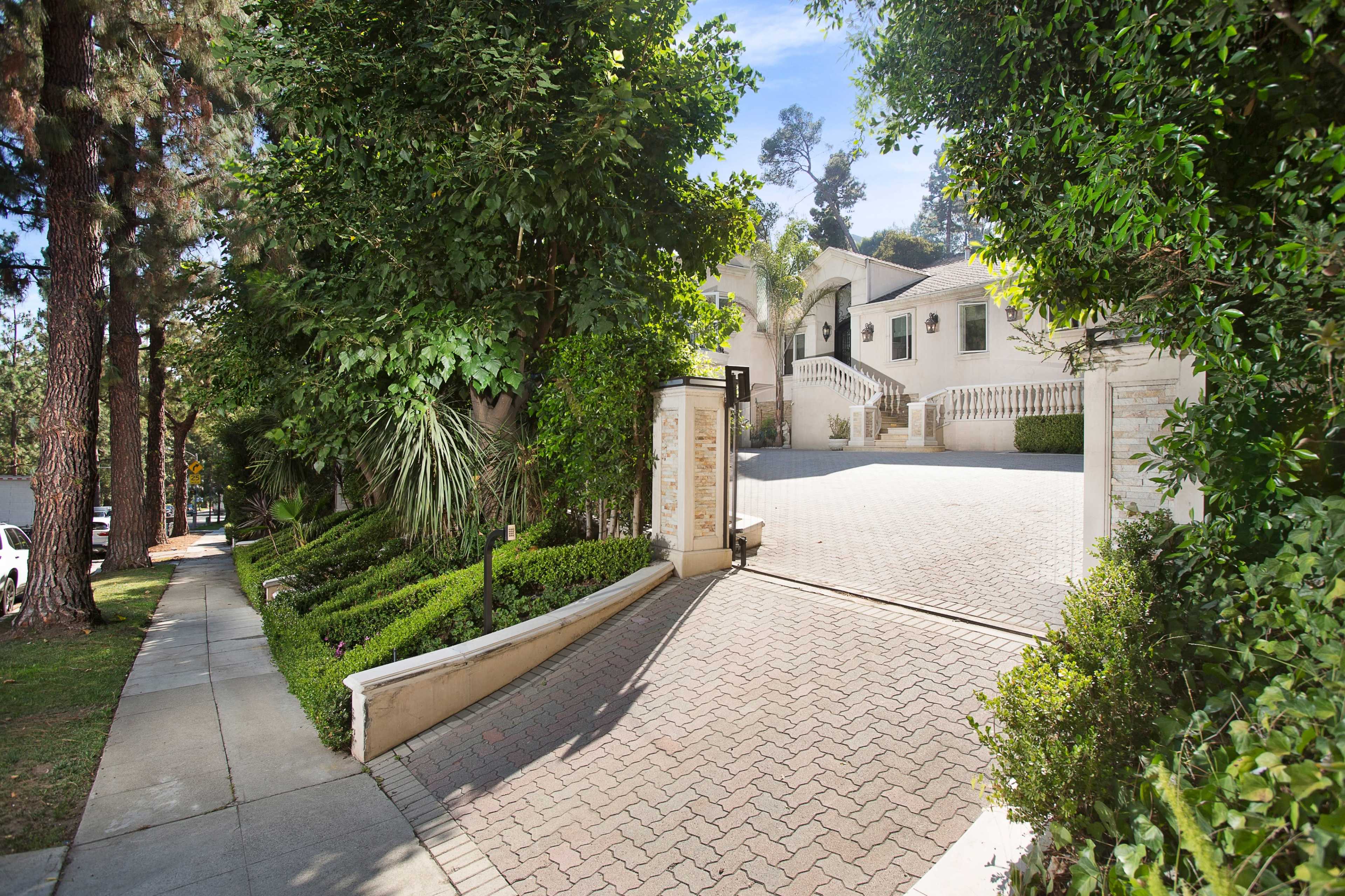 A driveway leading to a large, landscaped residential property behind a gated entrance, with trees lining the path.