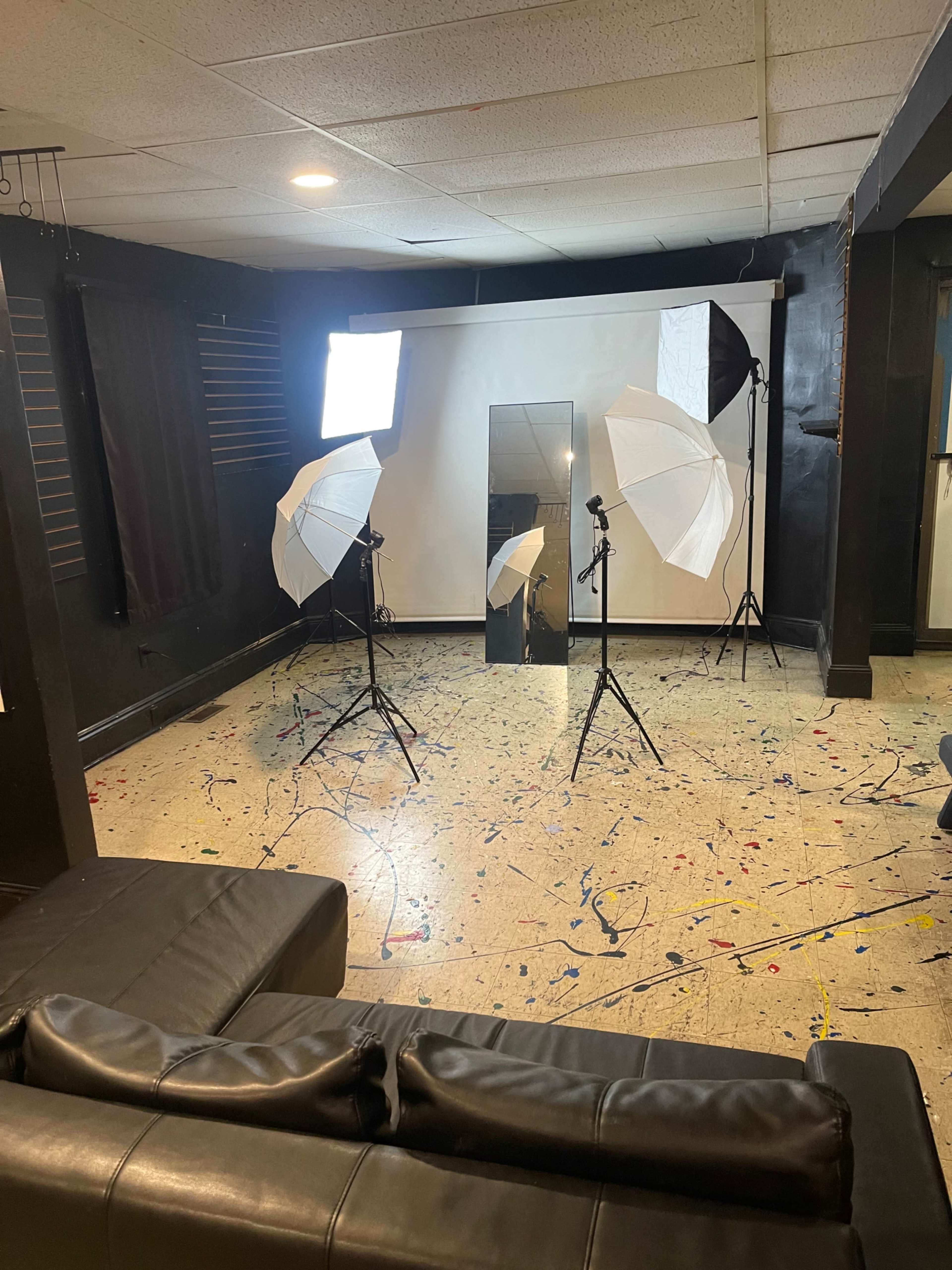 The image shows a photography studio setup with two softbox lights, a large mirror, and a white backdrop in an otherwise empty room featuring a colorful, splattered floor.