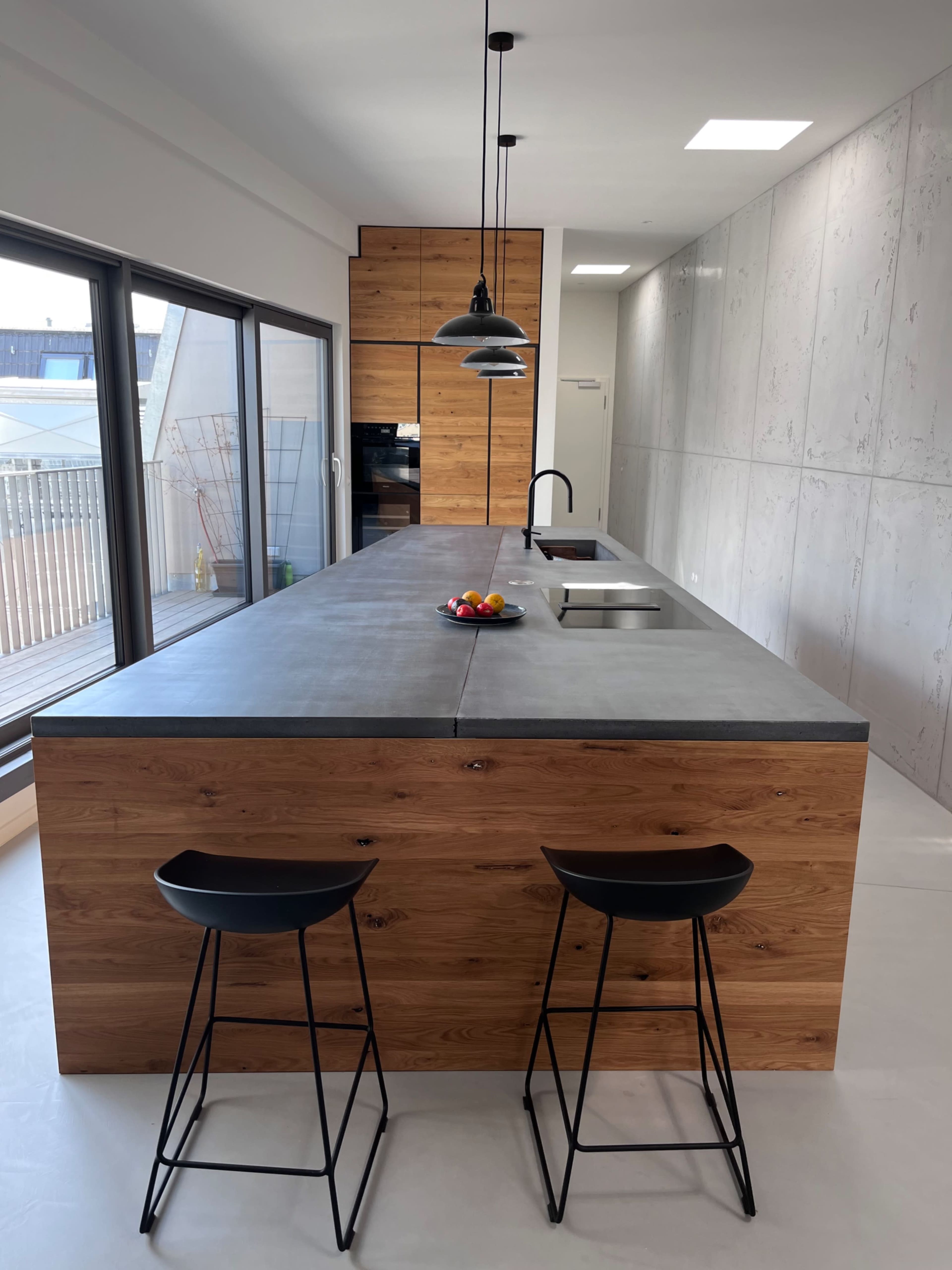 The image shows a modern kitchen with a large concrete countertop, wooden cabinetry, and two black stools.