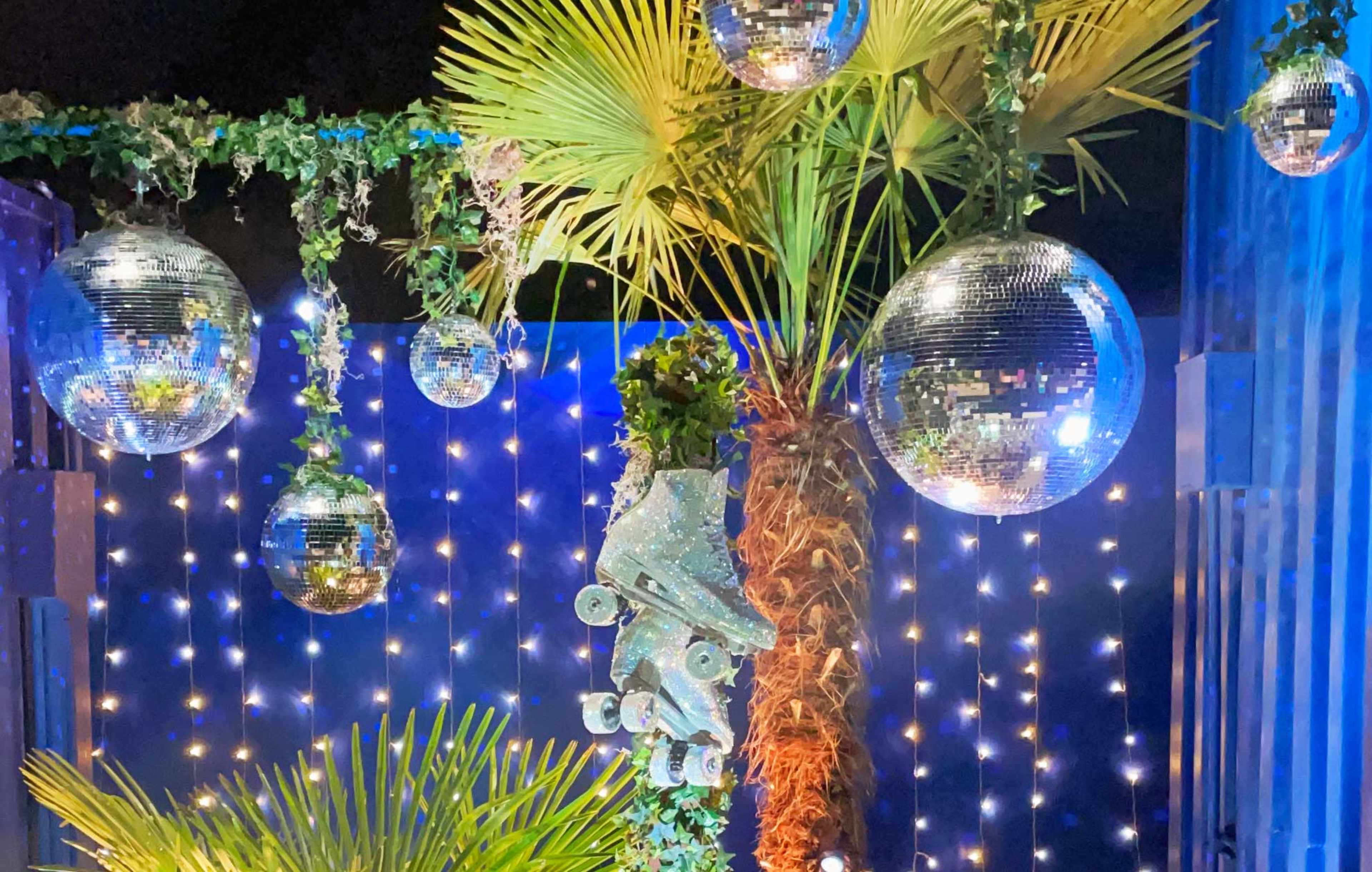 The scene features a vibrant setting adorned with hanging disco balls and a palm tree, illuminated by string lights.