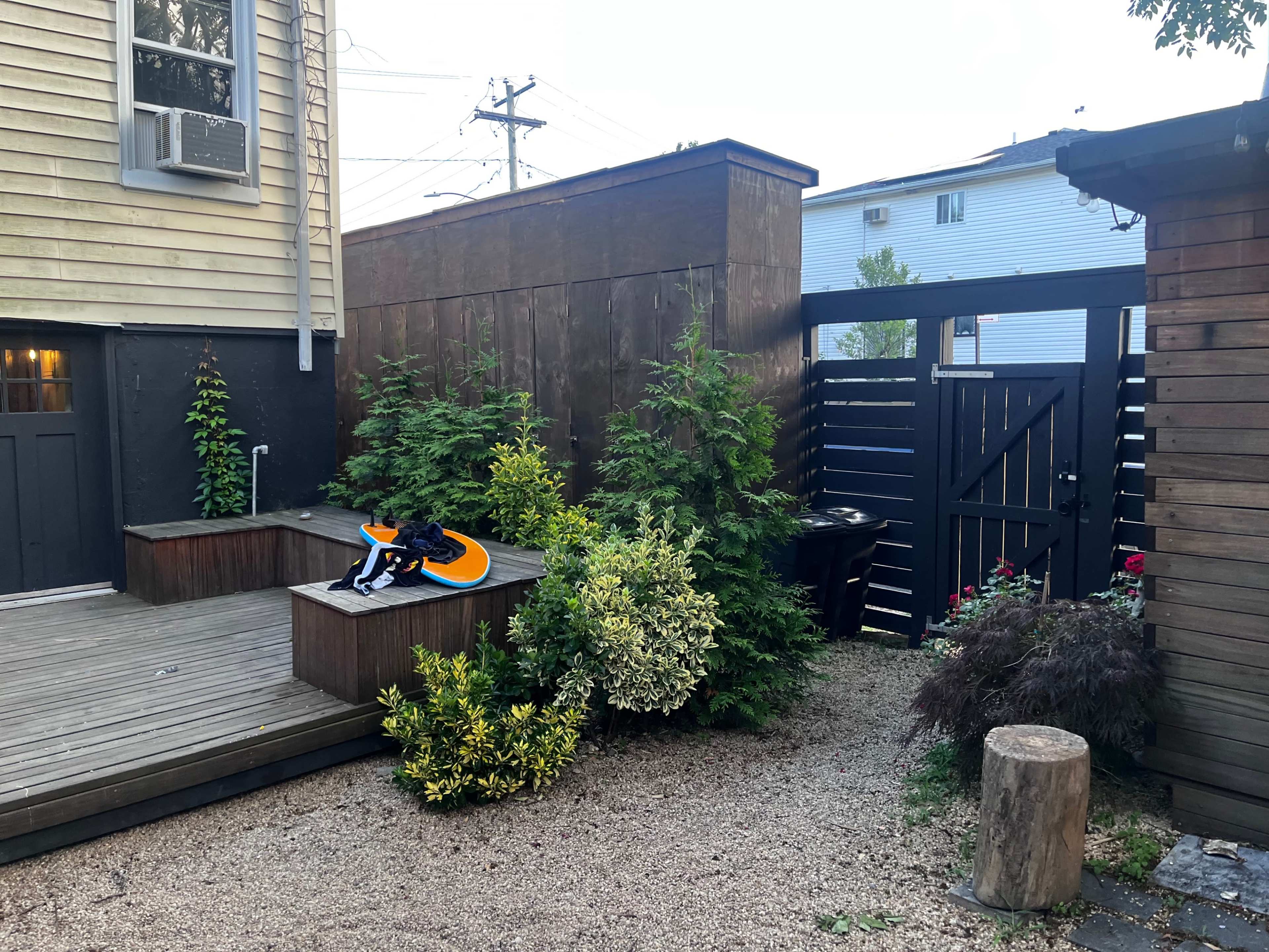 The image shows a wooden deck with a surfboard resting on it, surrounded by greenery and a fenced area with a gate in a backyard setting.
