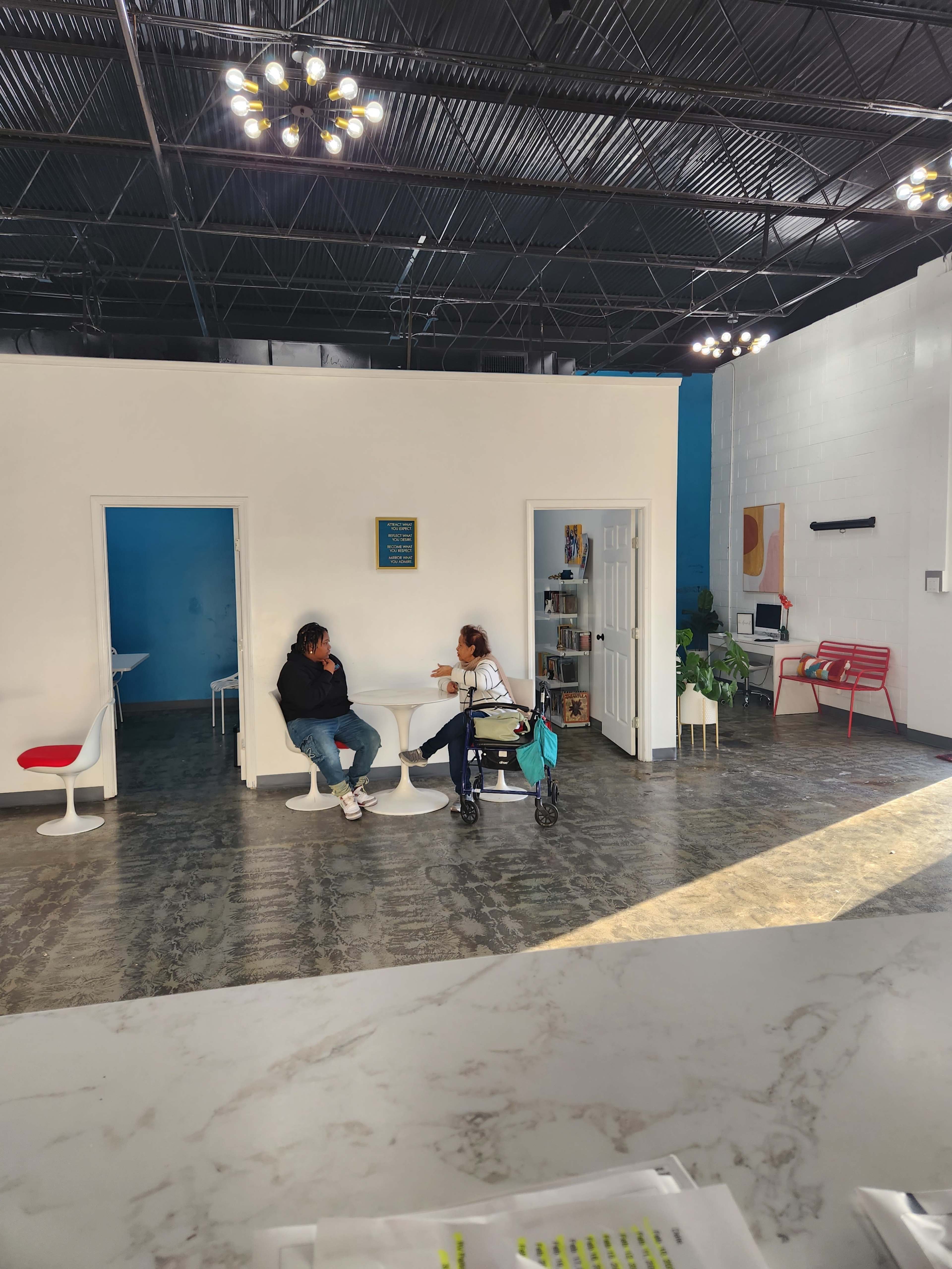 Two people are seated at a table in a bright, open room with modern furniture and large windows, while a third person is visible in a nearby doorway.