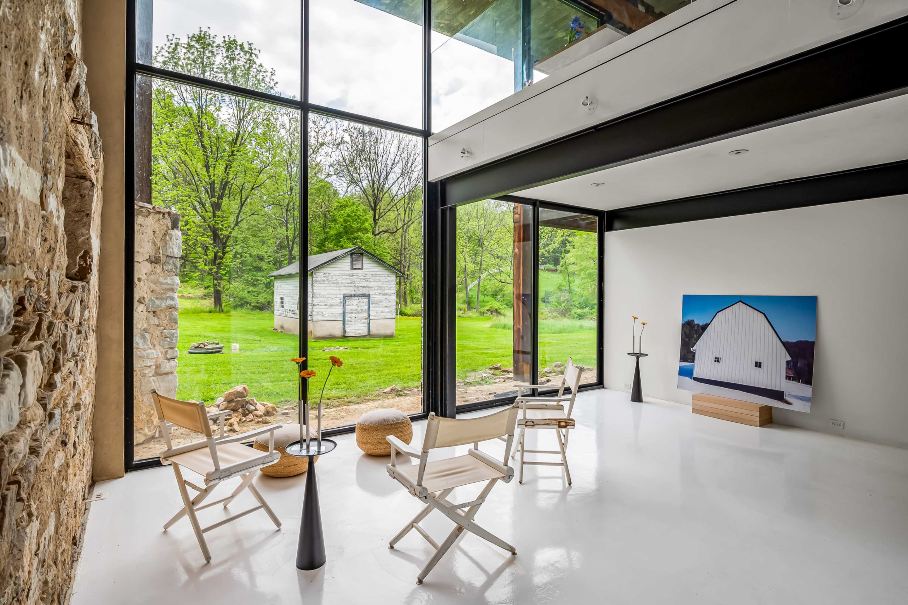 The image shows a modern interior space featuring white walls, large glass windows, and wooden beams, overlooking a green landscape with a small white shed.