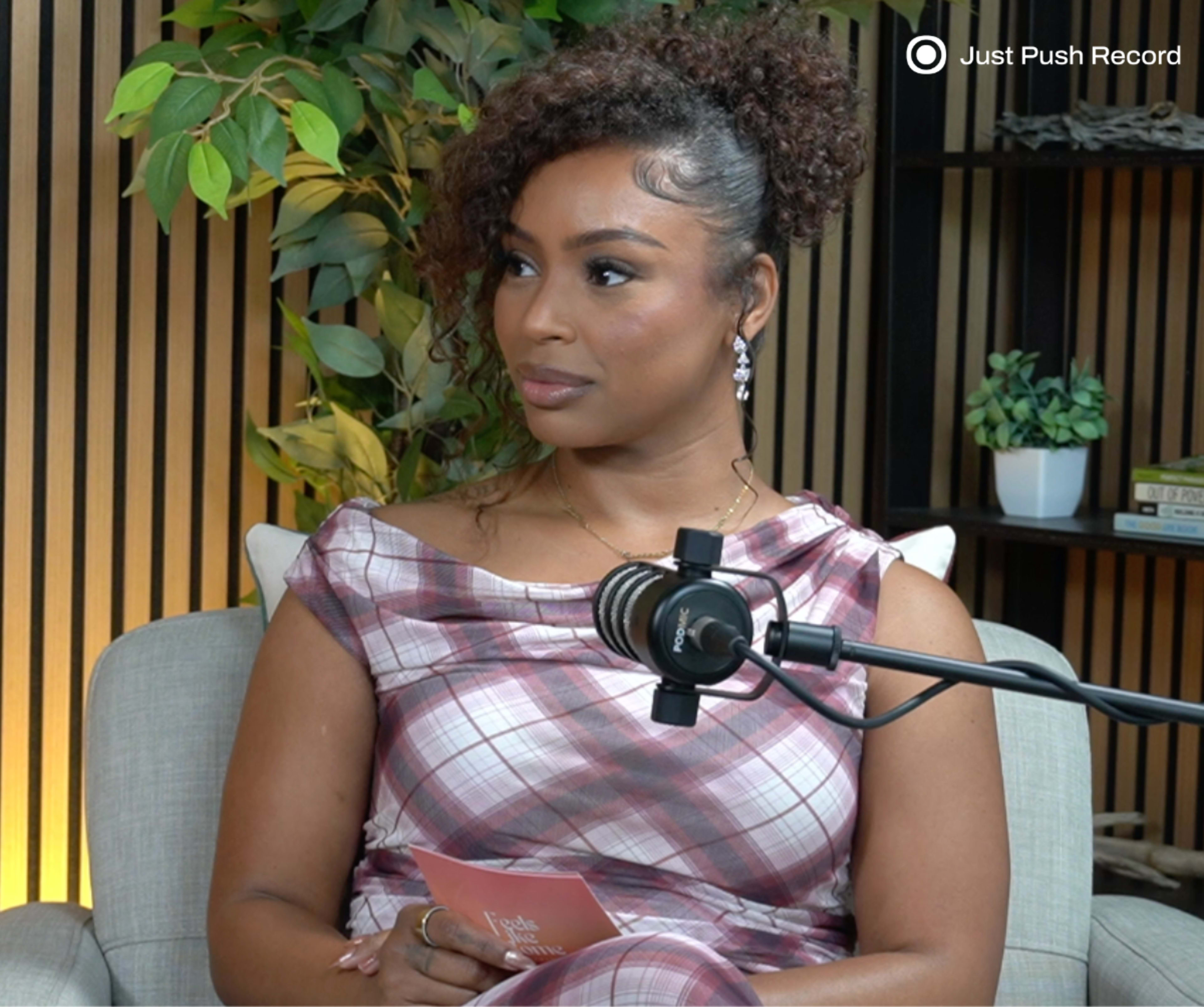 A woman with curly hair sits in front of a microphone and plants, holding a card and listening attentively.