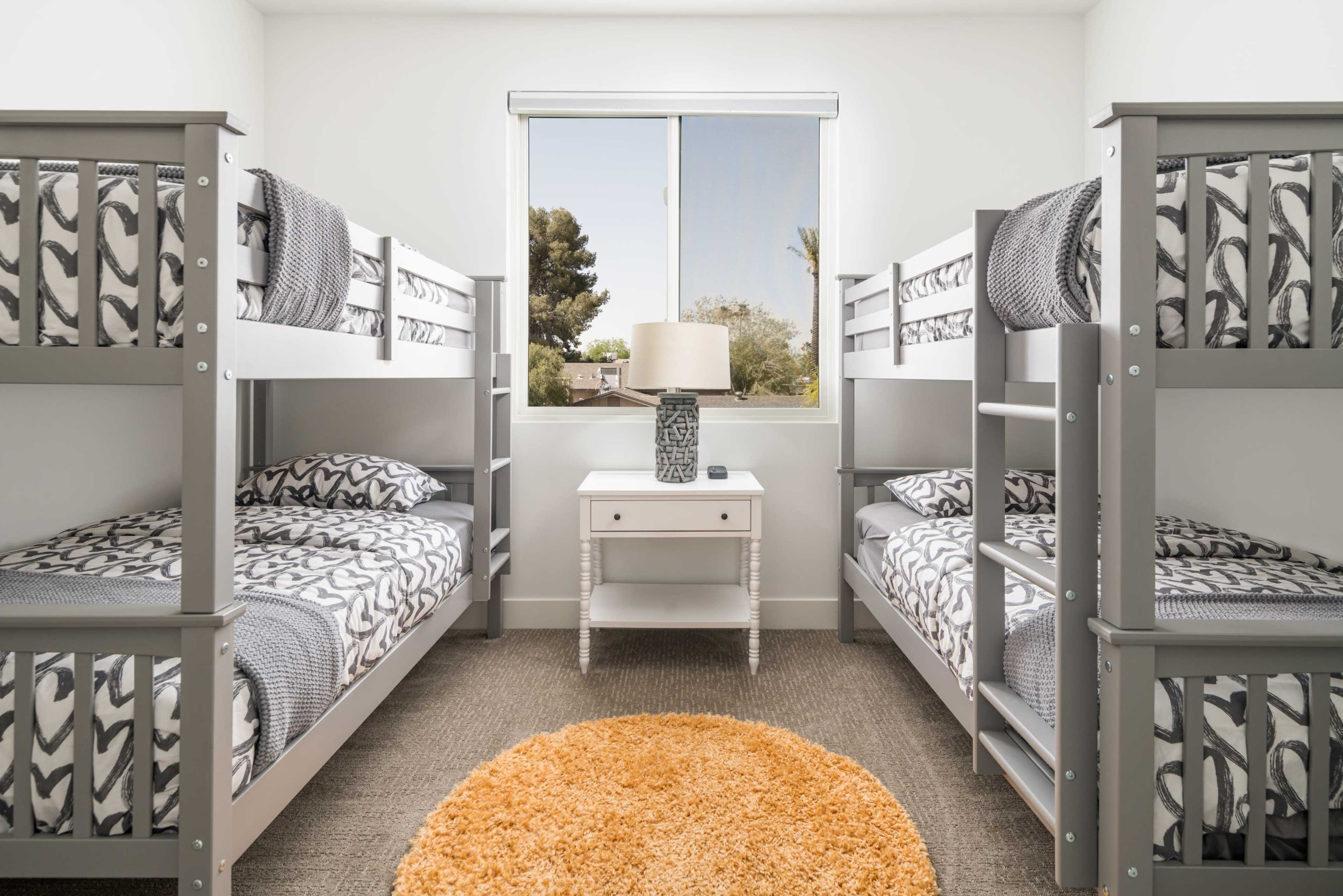 A bright bedroom with two sets of gray bunk beds, a bedside table with a lamp, and a round orange rug on the floor.