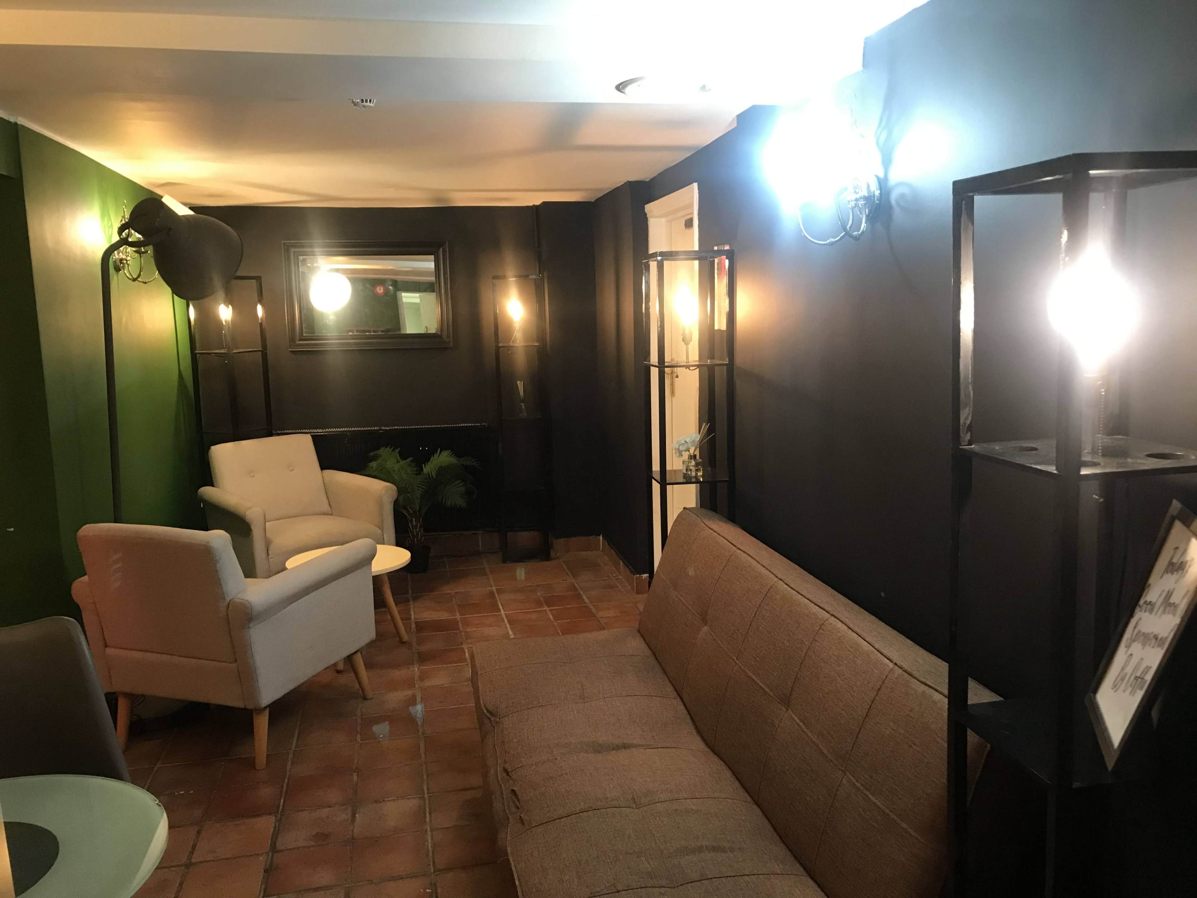 A cozy lounge area with a brown sofa, two light-colored chairs, a round table, and decorative light fixtures against a dark wall.