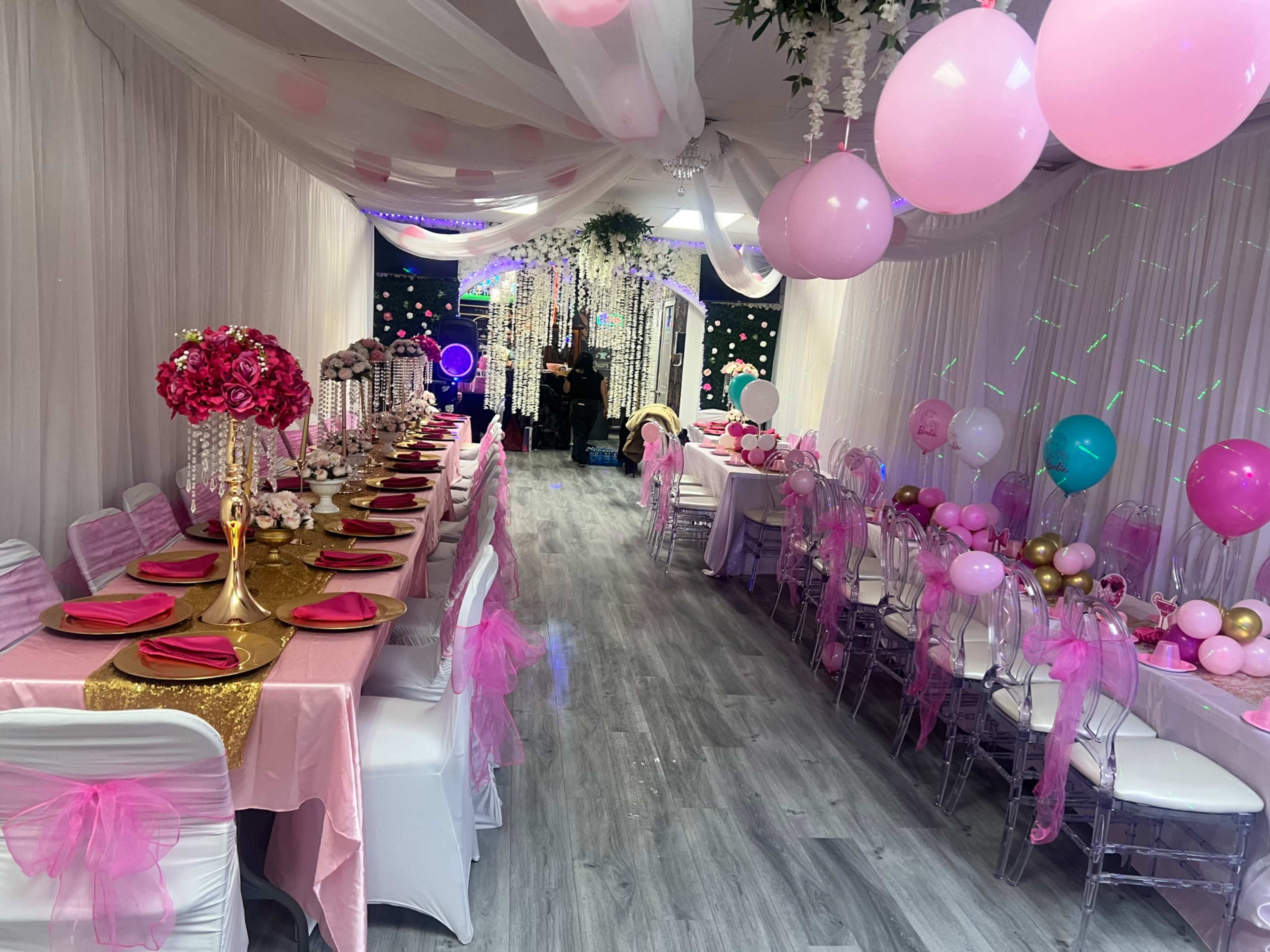 The image depicts a decorated event space featuring long tables set with pink tablecloths, white chairs adorned with ribbons, and various pink and gold balloons.