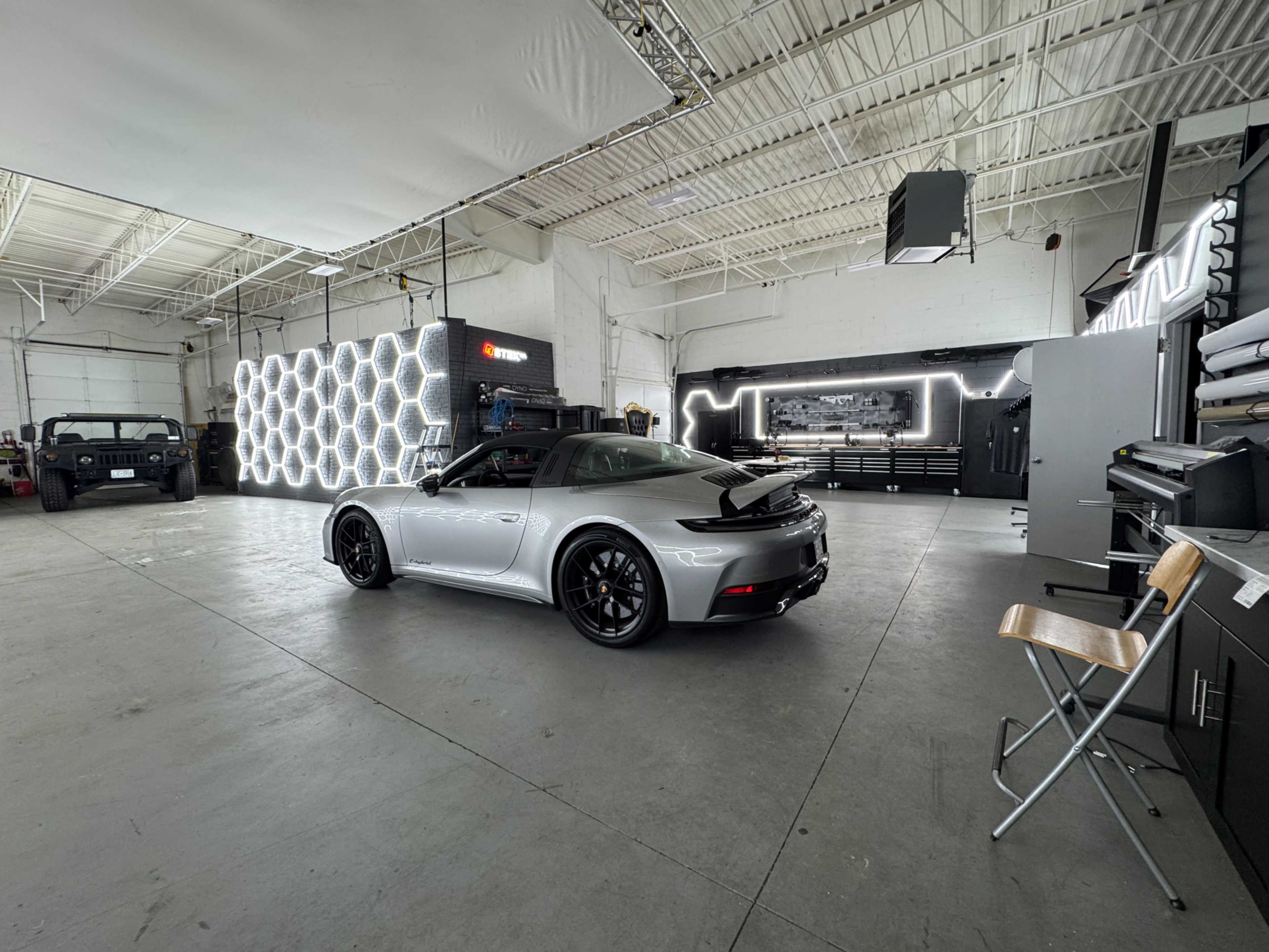 A silver sports car is parked in a spacious garage featuring a modern design with illuminated hexagonal wall panels and various equipment arranged in the background.