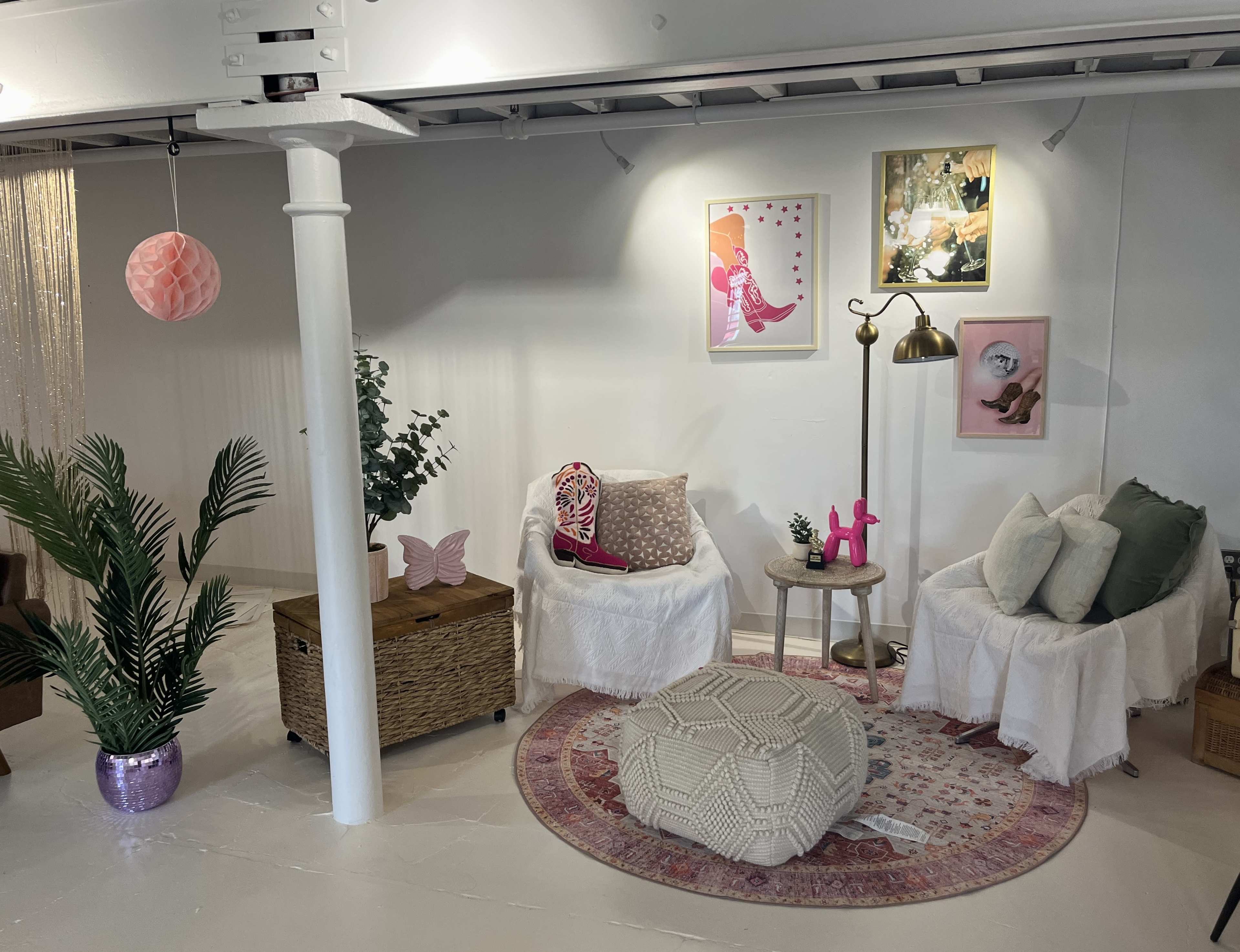 A cozy room corner with two armchairs, a small table, decorative artwork on the walls, a large pouf, and a potted plant.