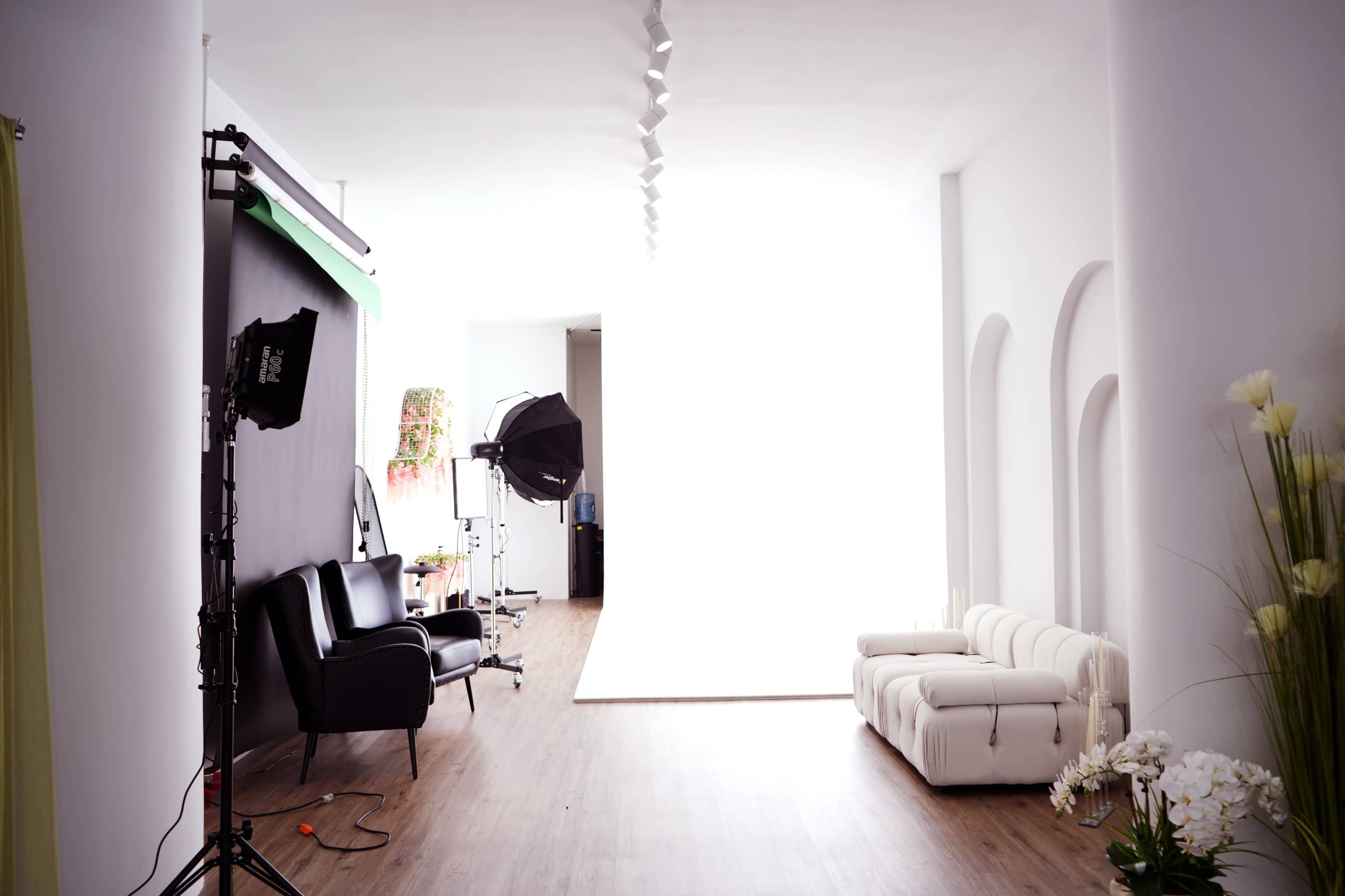 The image shows a spacious photography studio with a white backdrop, a black sofa, a white couch, and lighting equipment set up.