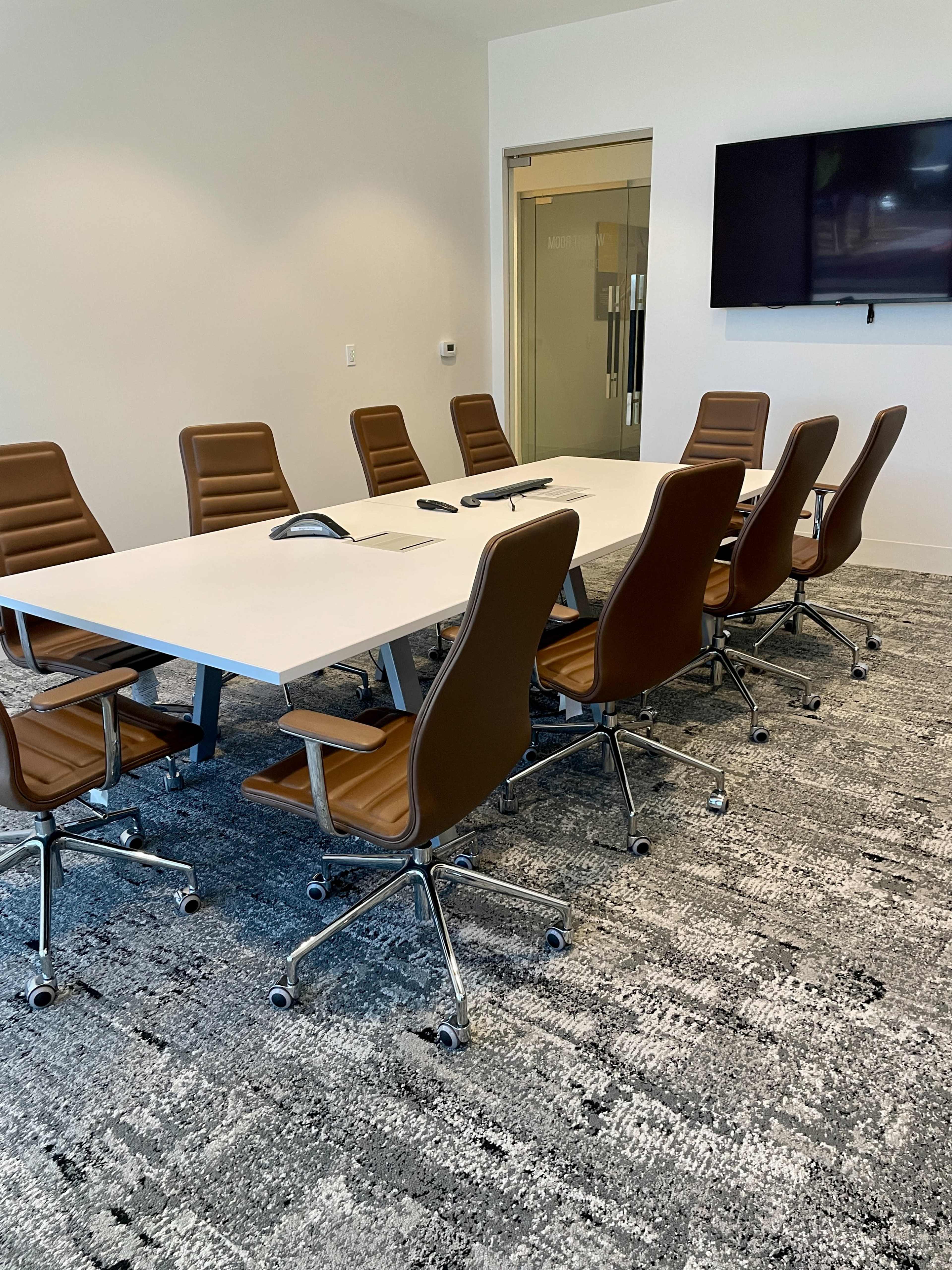 A modern conference room features a large white table surrounded by ergonomic brown chairs, with a television mounted on the wall and sliding glass doors.