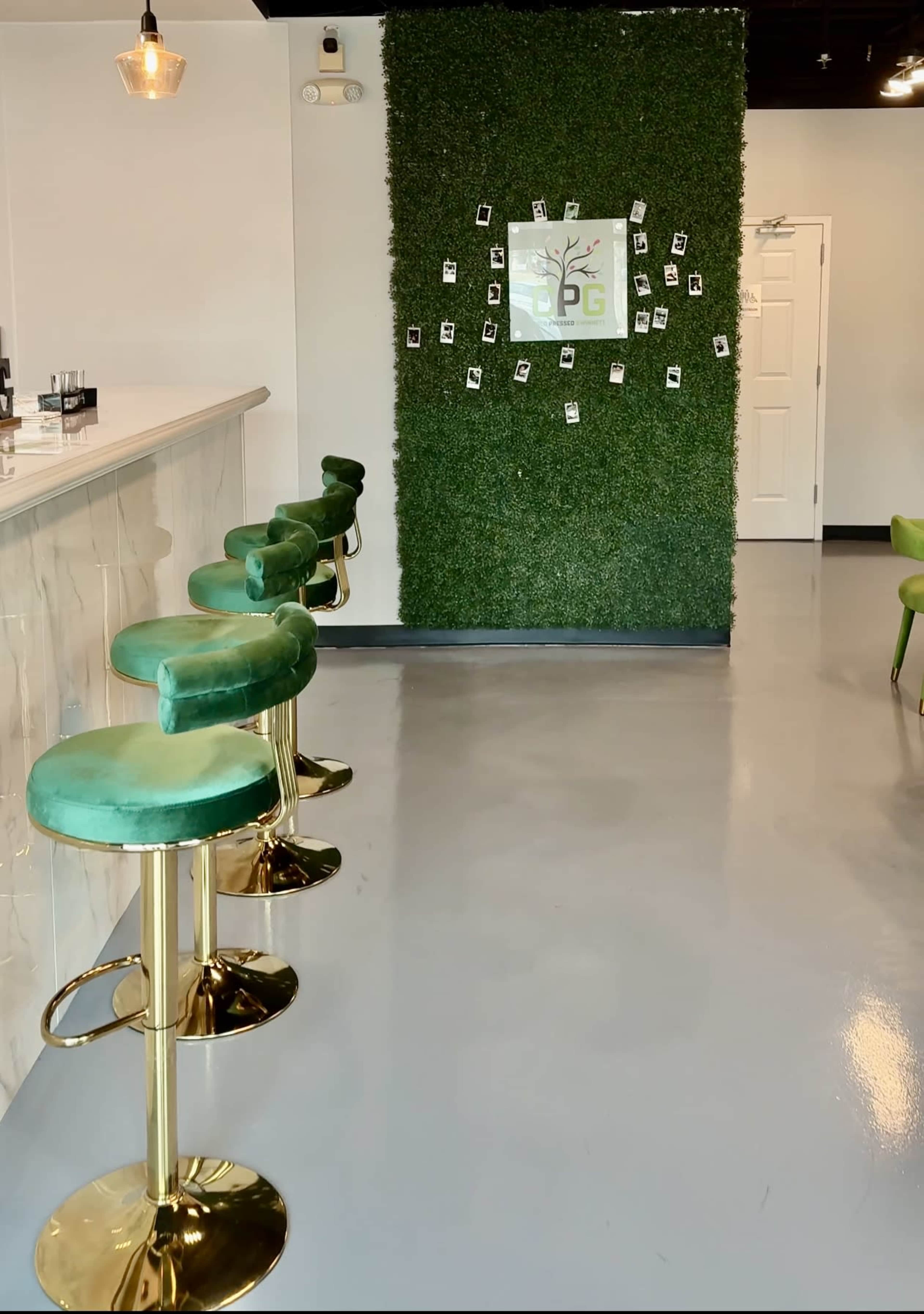The image shows a modern café interior with green bar stools lined up at a counter, a wall covered in artificial greenery, and decorative photos displayed.