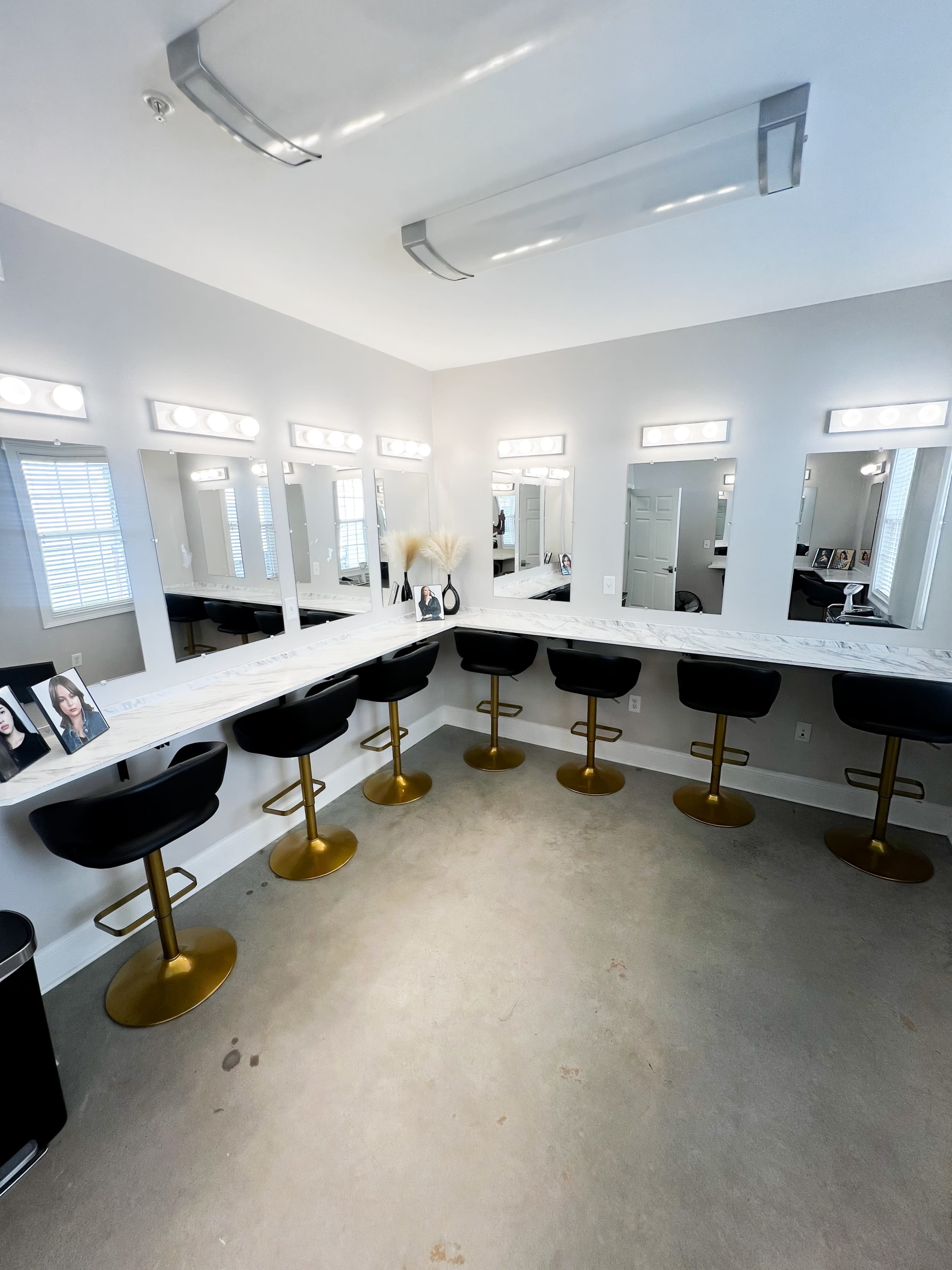 The image shows a modern makeup studio featuring a long marble countertop with five black barstools and illuminated mirrors arranged along the wall.