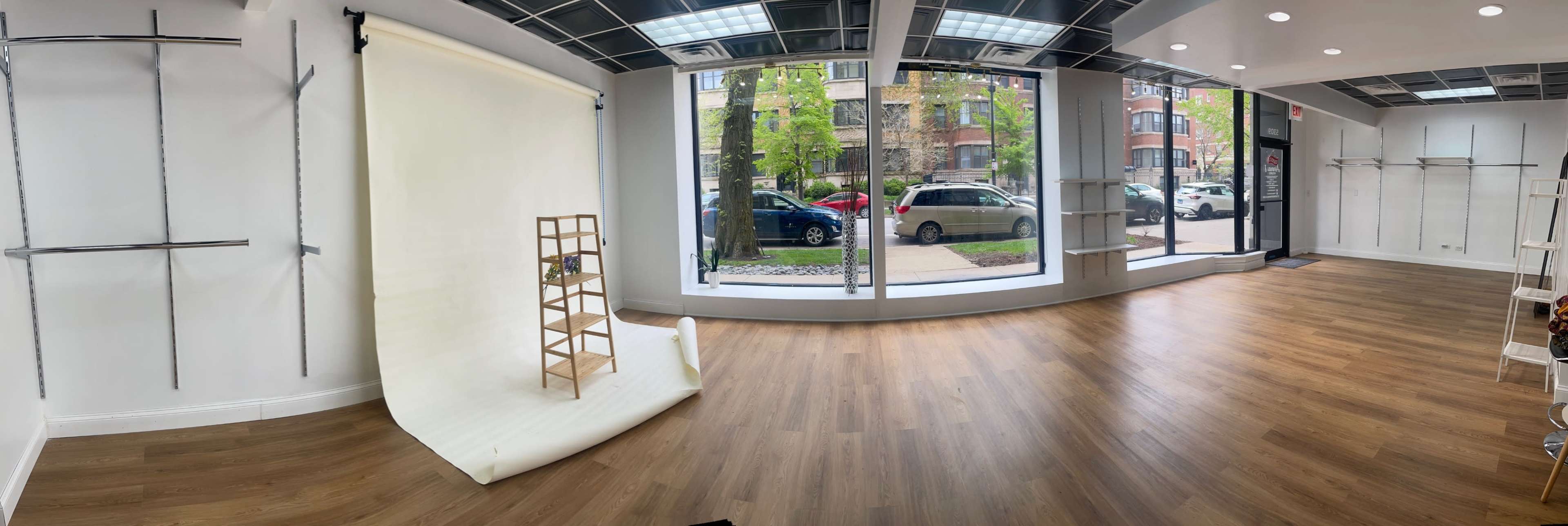 The image depicts an empty retail space featuring a light-colored backdrop on one side, wooden flooring, and large windows displaying outside scenery and parked cars.