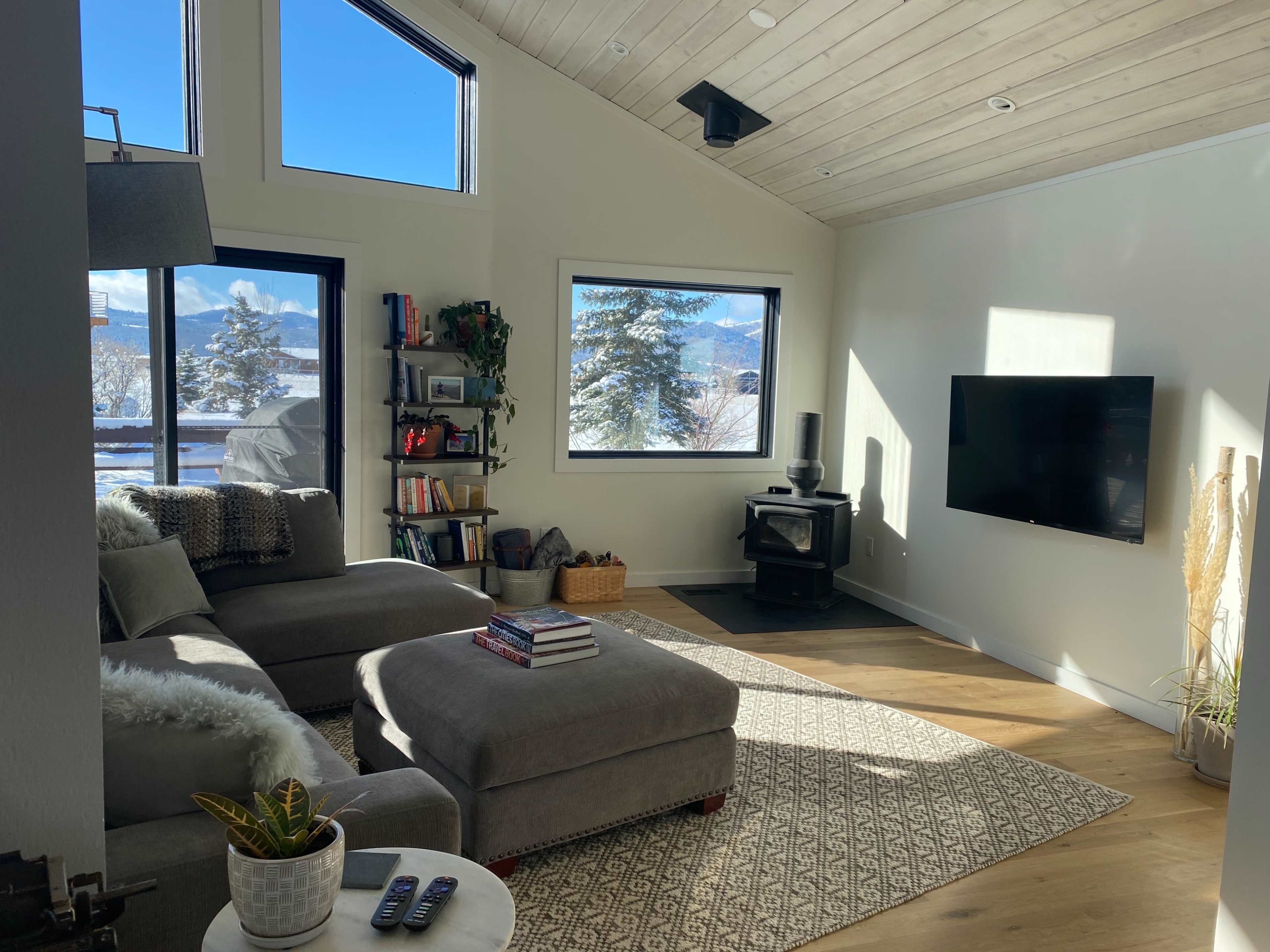 The living room features a gray sectional sofa, a wall-mounted television, a wood stove, and large windows displaying a view of mountains and trees.