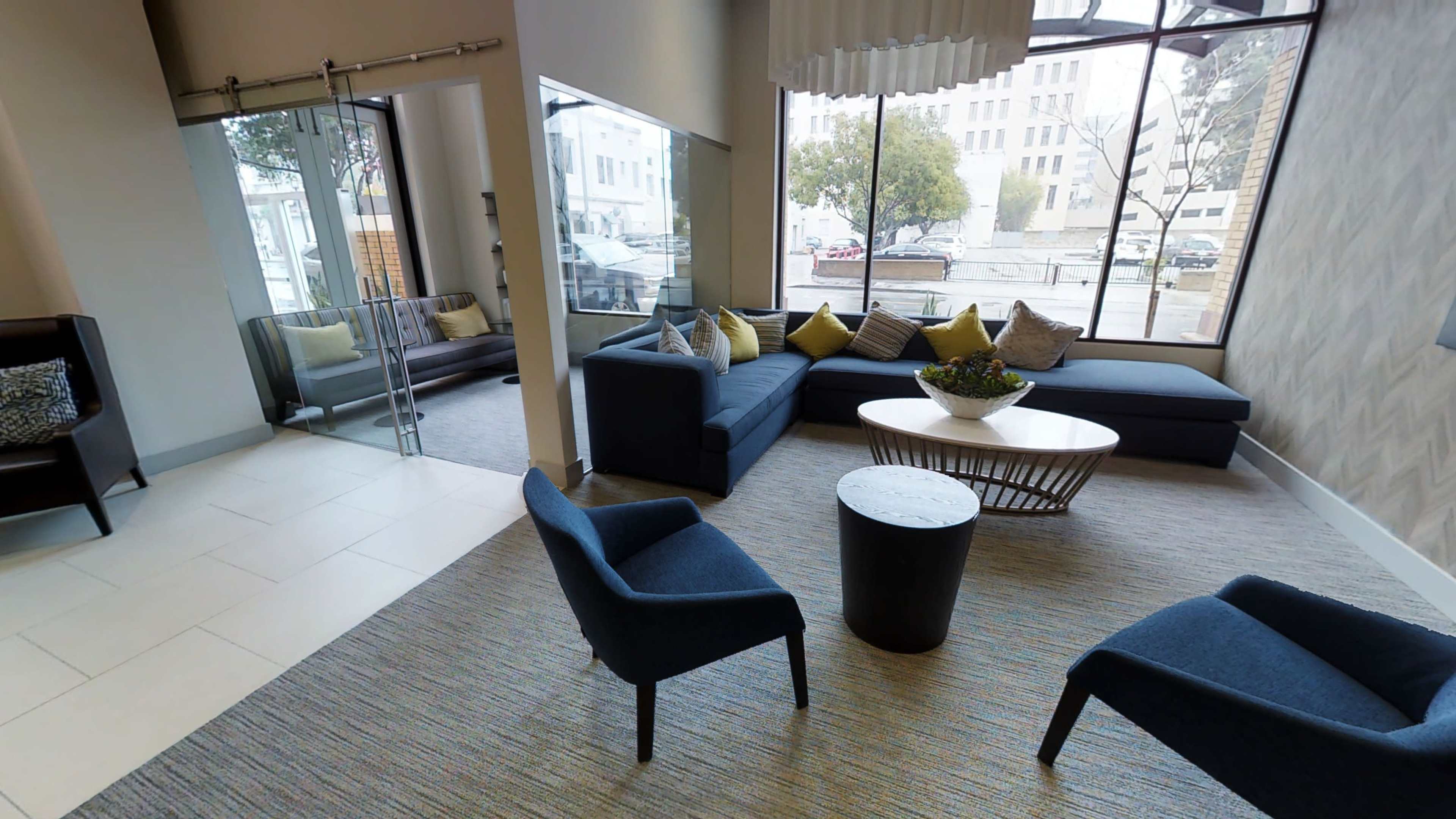 The image shows a modern lobby area with a large sofa, two blue chairs, a round table, and large windows letting in natural light.